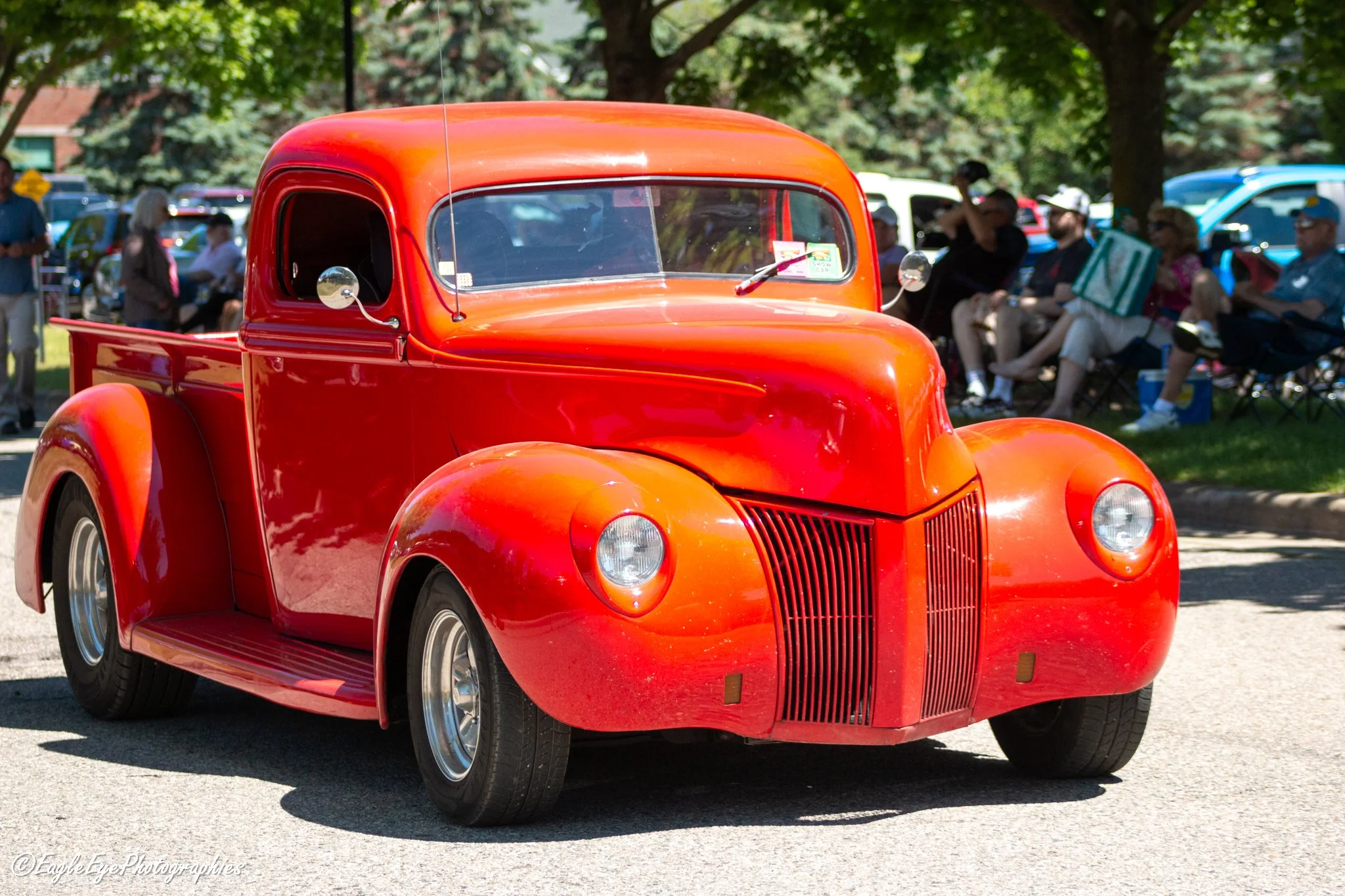 A vintage red pickup truck parked outdoors during a car show, with people seated and walking in the background, surrounded by trees.
