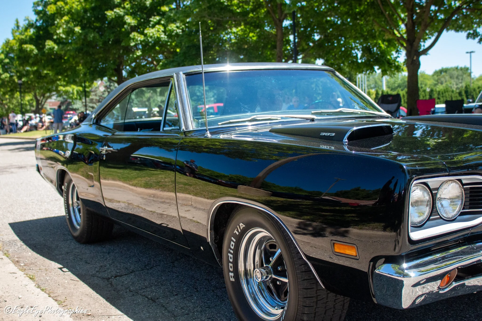 Black classic muscle car with chrome accents parked outdoors under green trees on a clear sunny day.