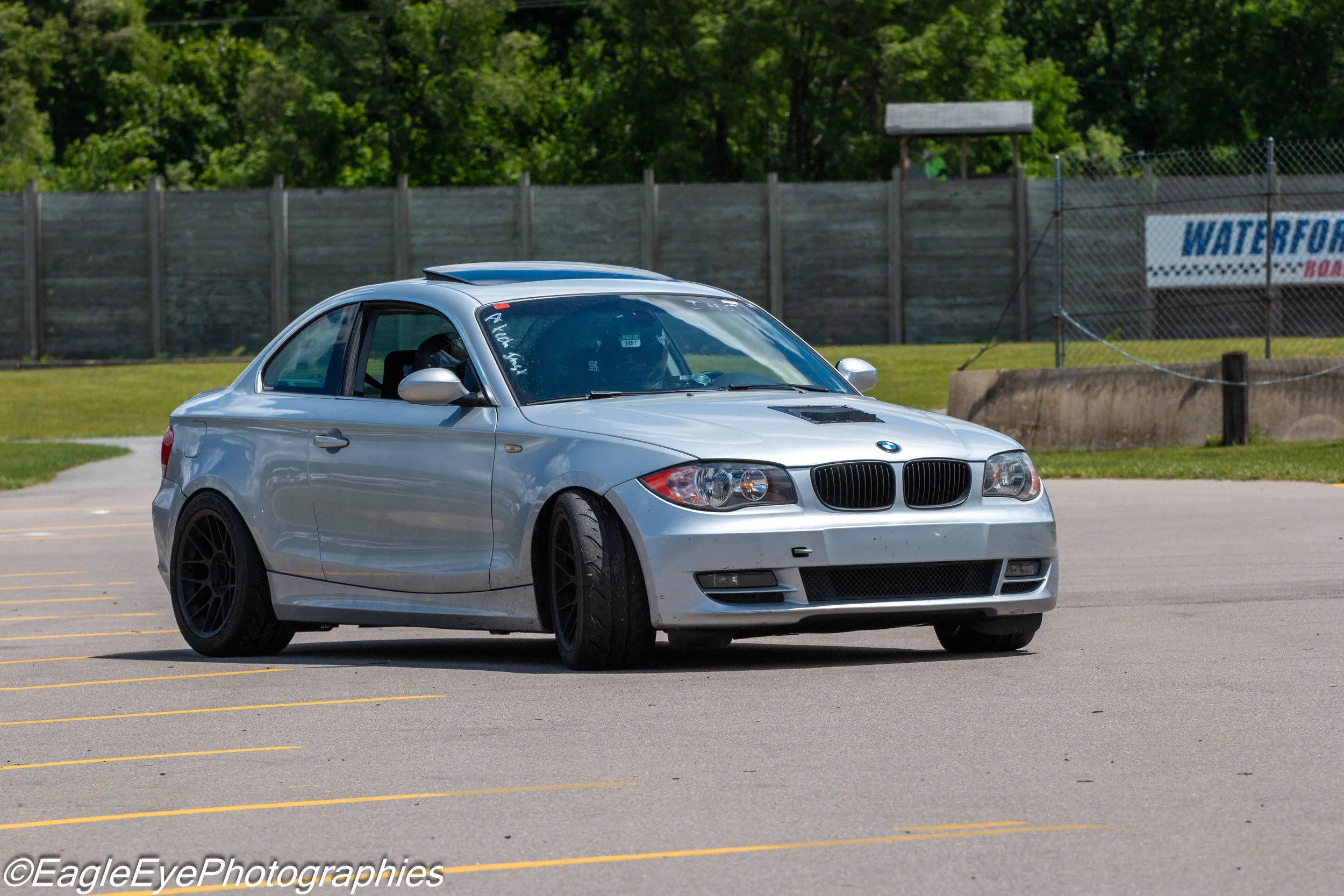 A silver BMW car parked in an outdoor lot with a wooden fence and trees in the background.