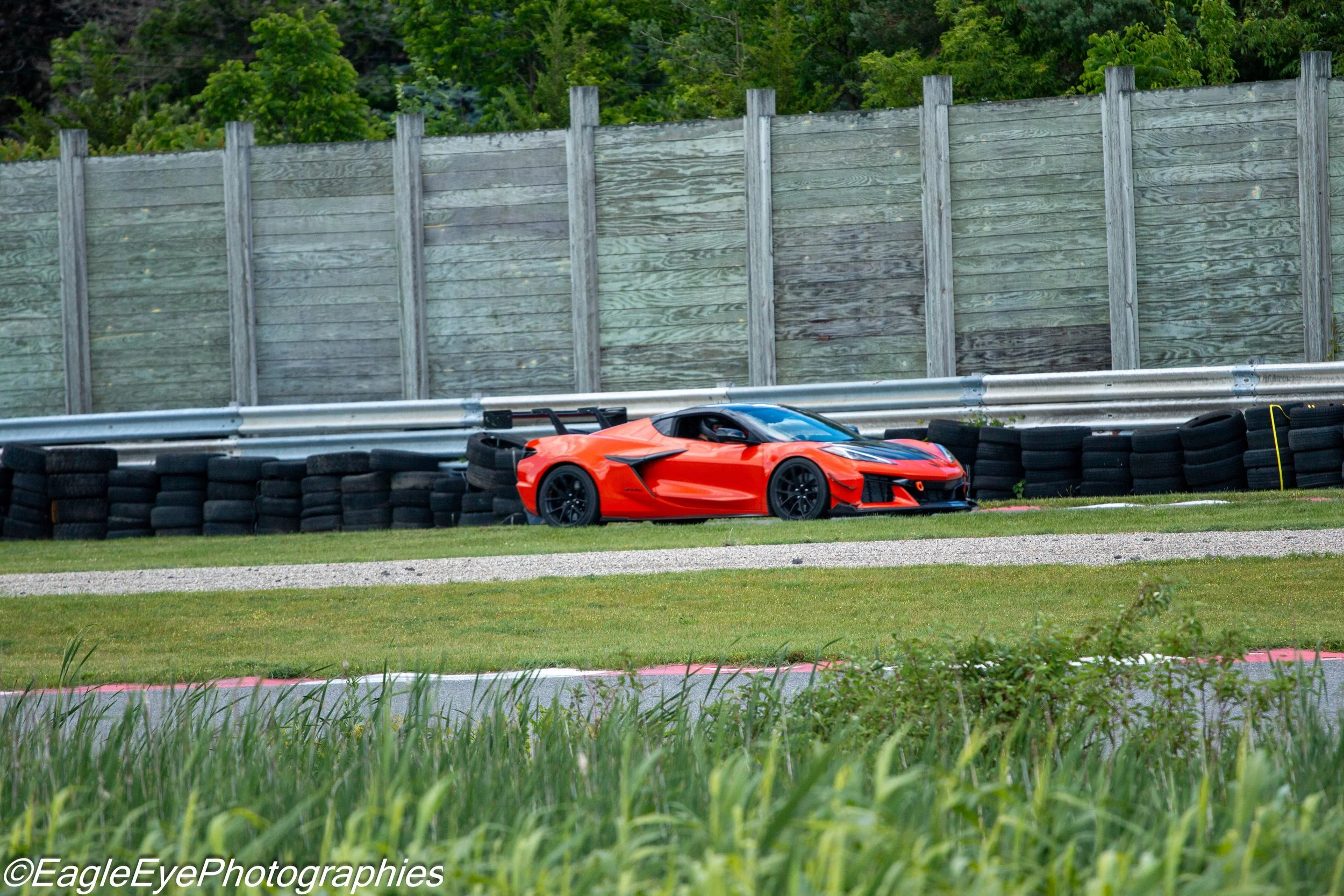 A bright orange sports car on a racetrack with grass in the foreground, a tire barrier, metal guardrail, and a wooden fence with green trees behind it.
