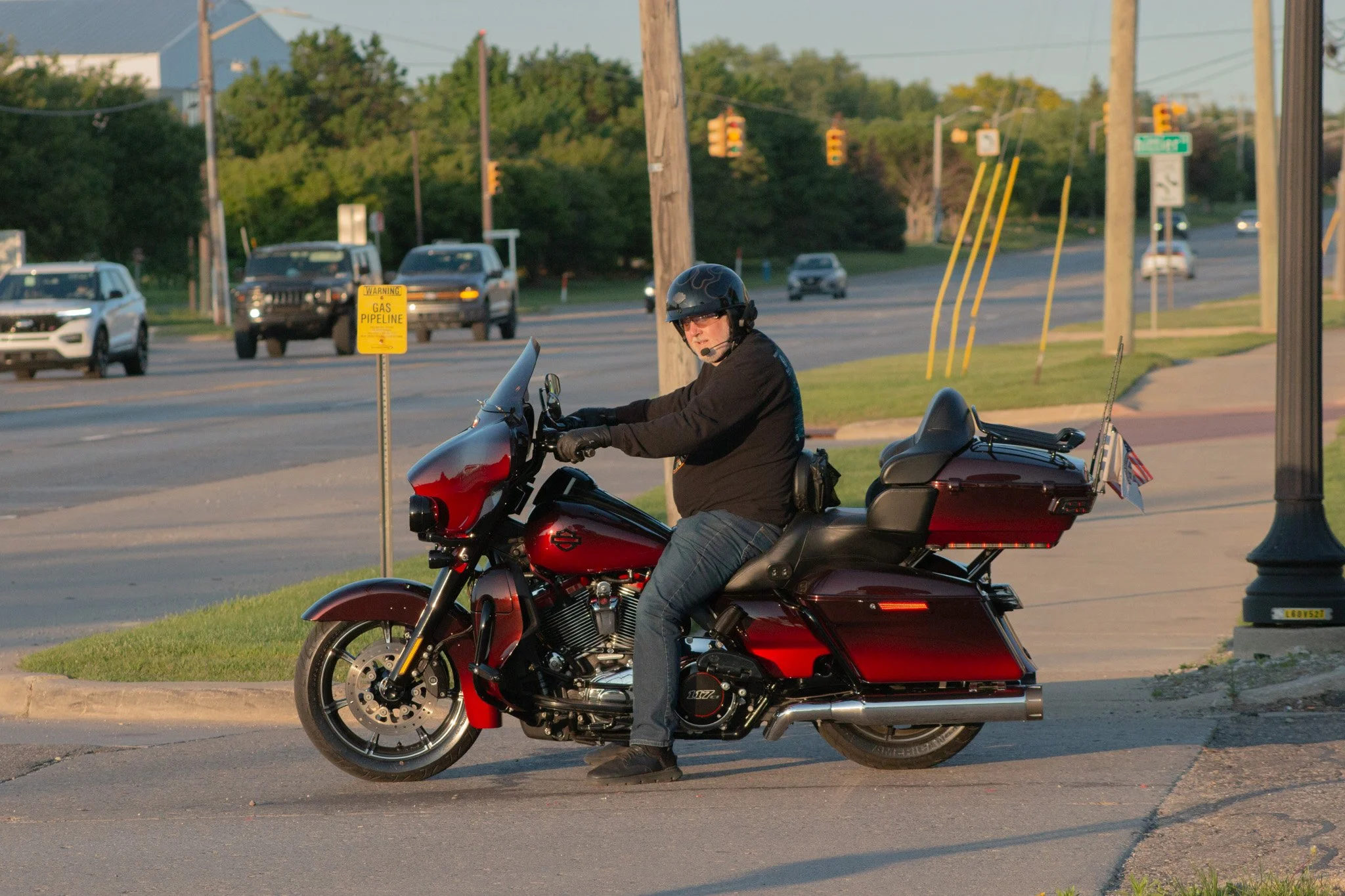 A man wearing a black helmet and glasses riding a red motorcycle along a street. The motorcycle has a large back storage compartment with a small American flag attached. There is a yellow warning sign about a gas pipeline nearby. Traffic is visible i