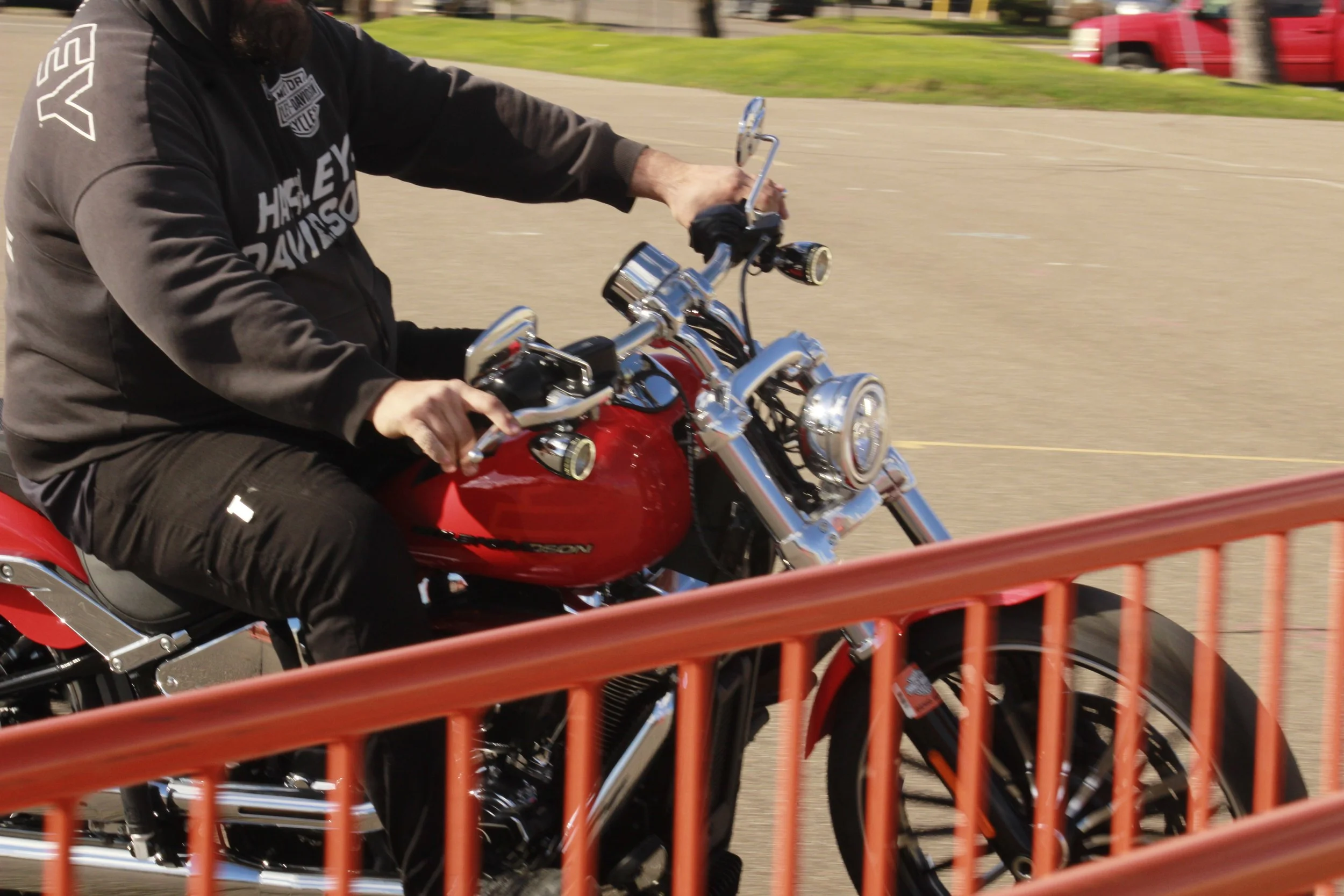 A person riding a red Harley Davidson motorcycle on a city street, captured in motion with orange railings in foreground and a grassy area with a red vehicle in the background.