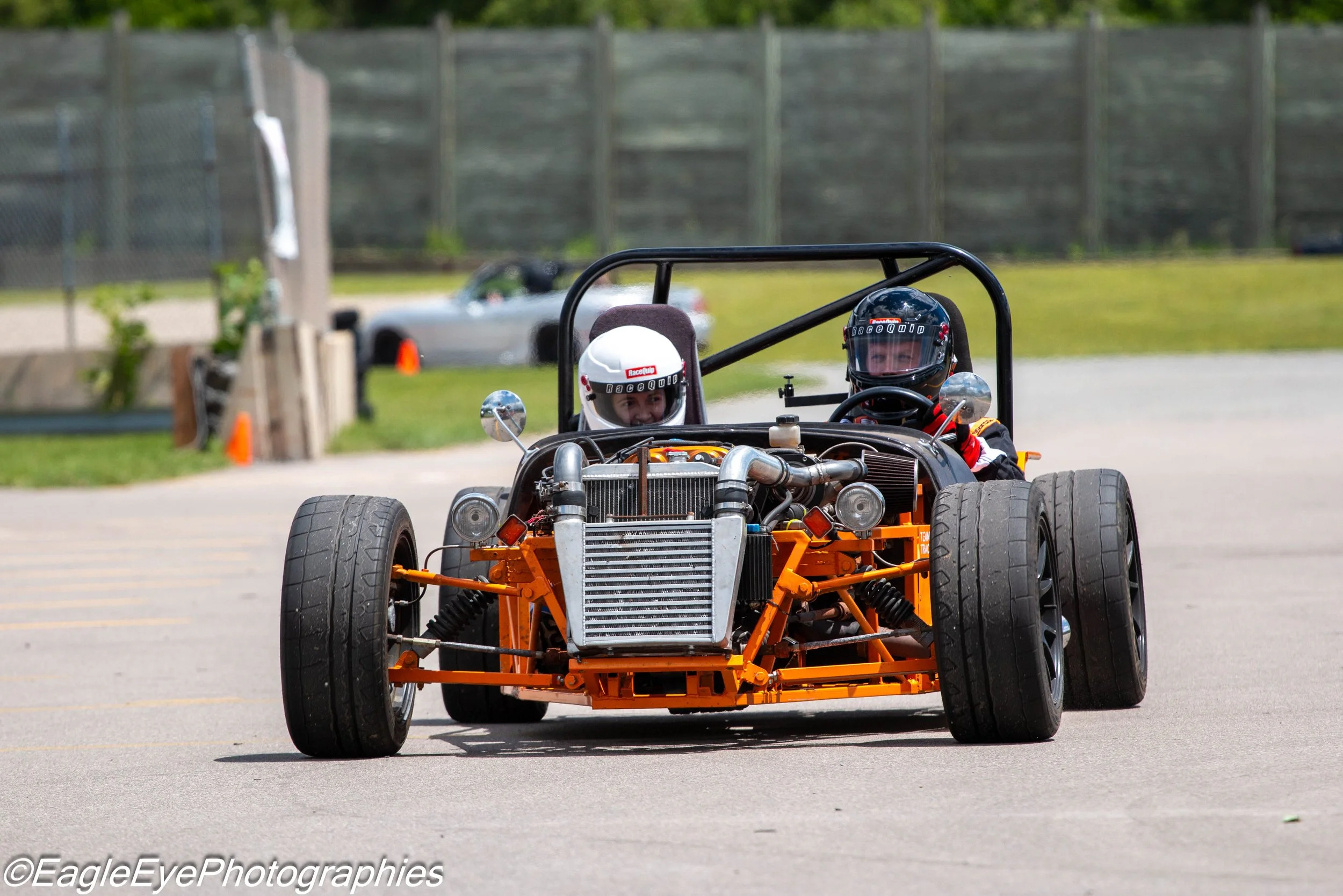 Two people in racing helmets driving a small, open-wheel race car on a paved track, with a blurred background of a fence and a car.
