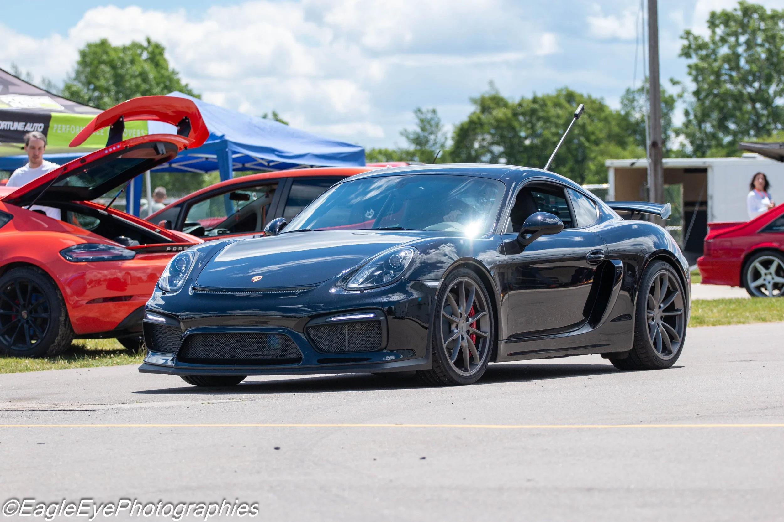 A black sports car parked outdoors with other colorful cars and tents in the background, at what appears to be a car show.