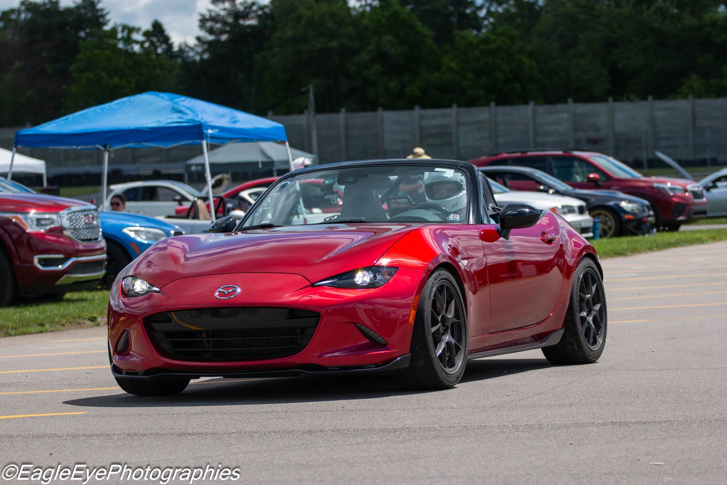 Red Mazda convertible sports car parked in a parking lot with other cars and tents in the background.