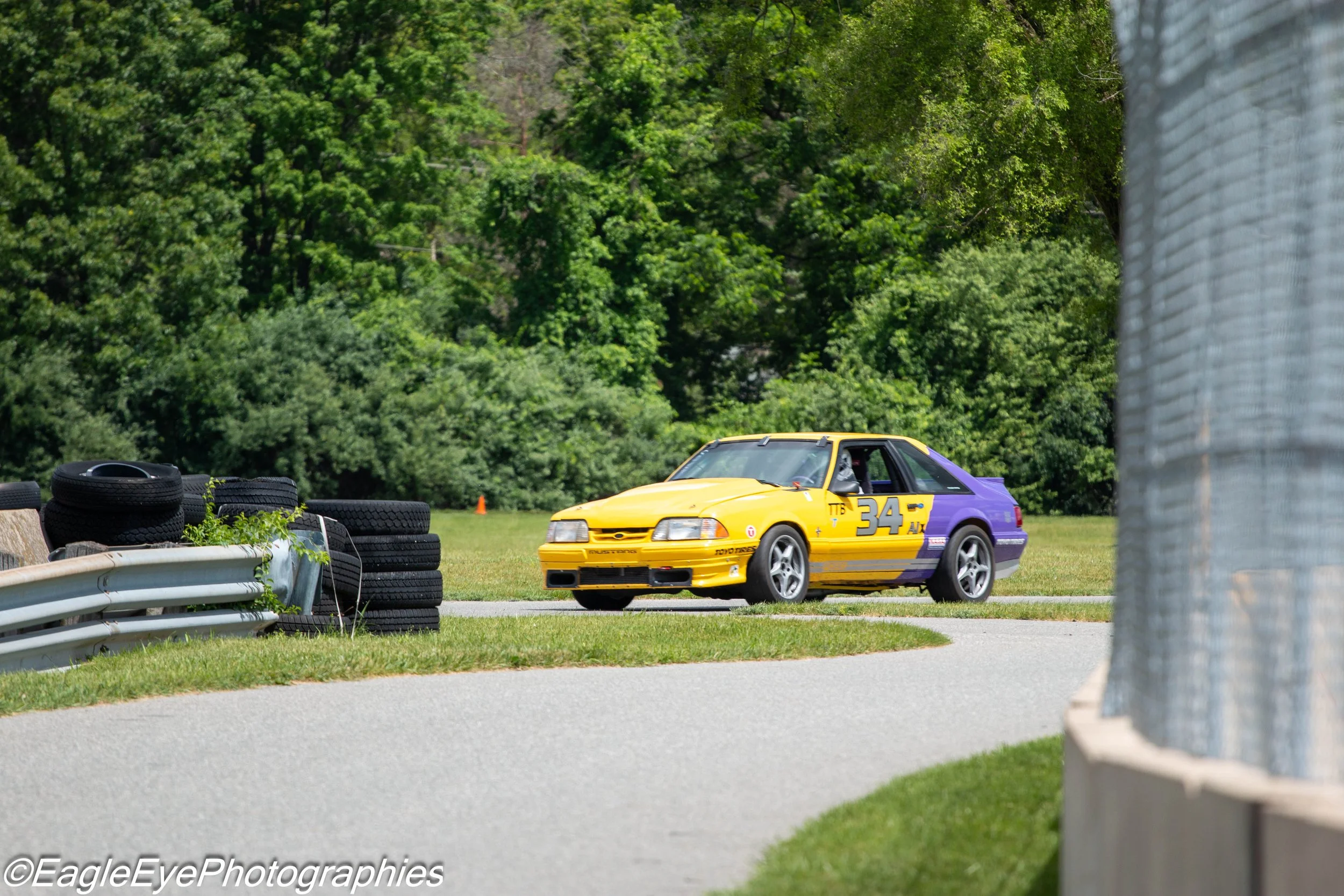 A yellow and purple racing car with the number 34 on its side, parked on a grassy area near a stack of tires and a metal guardrail, with trees in the background.