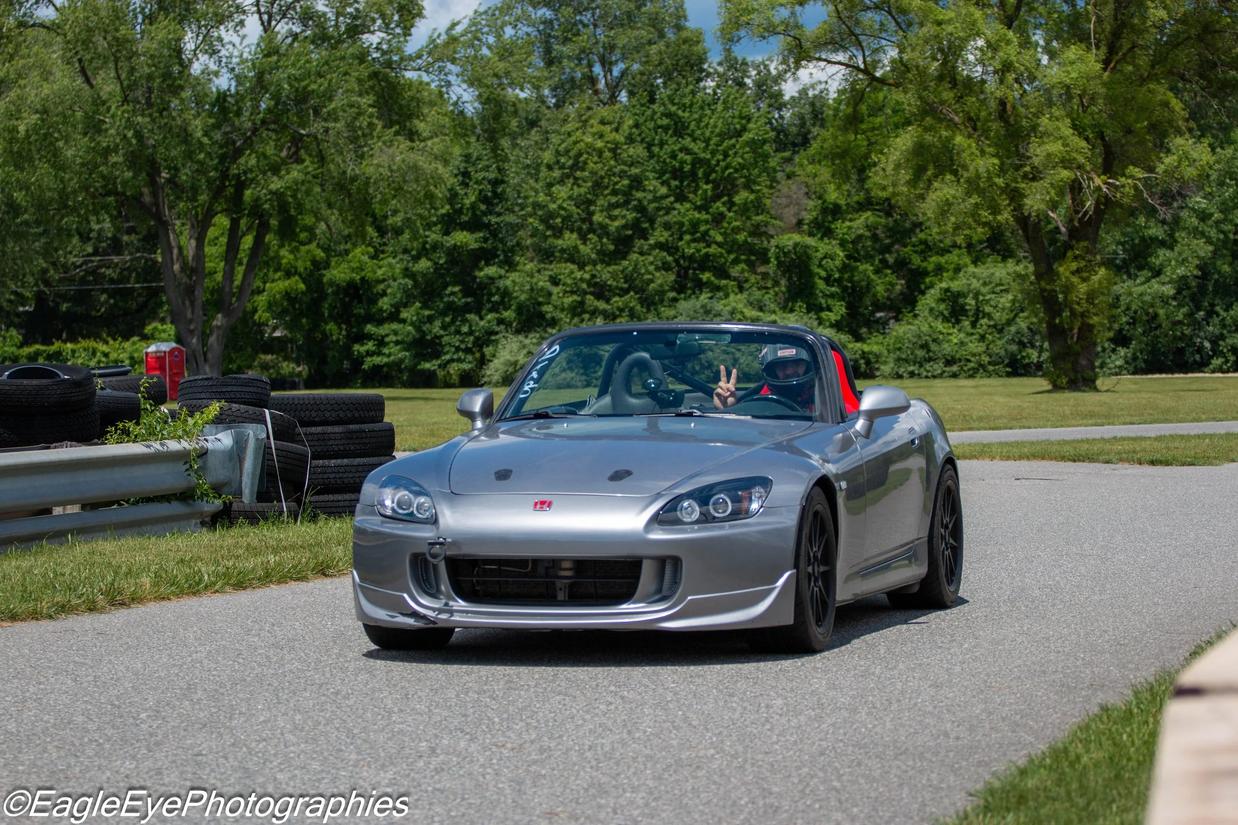 A silver sports car with a person in a helmet and racing gear making a peace sign through the windshield, parked on the side of a road near a stack of tires and lush green trees.