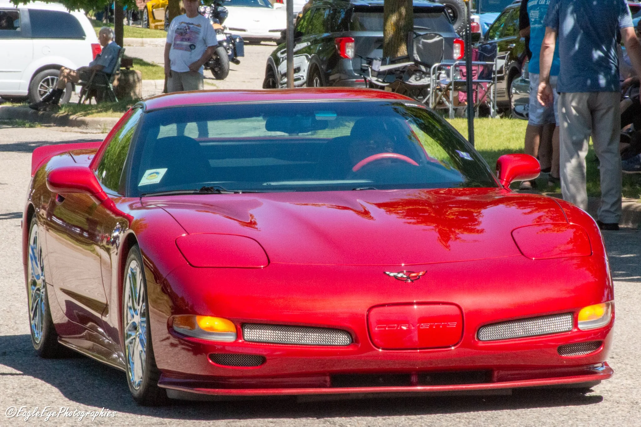 A red Chevrolet Corvette sports car parked outdoors with people and vehicles in the background at a sunny event.