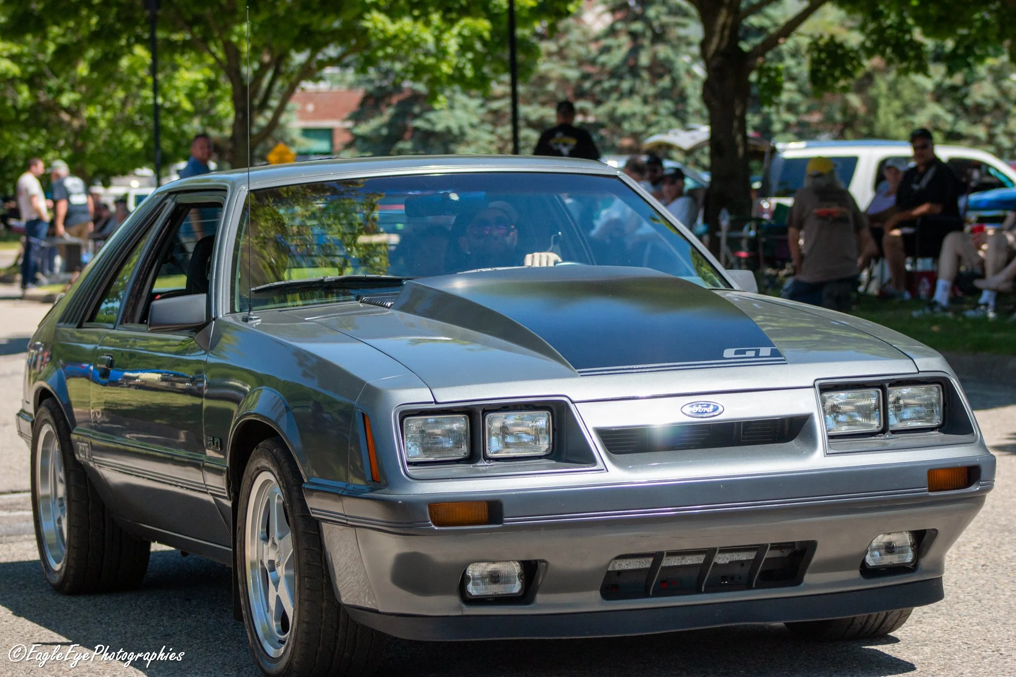 A silver vintage sports car parked outdoors at a car show, with a crowd of people, trees, and other vehicles in the background.