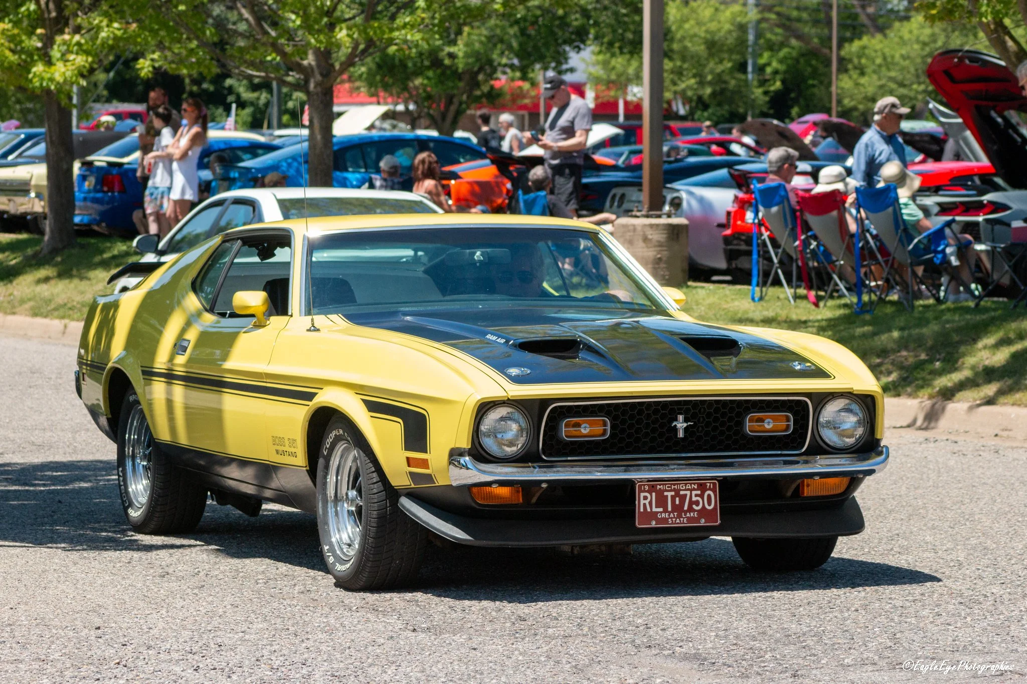 A yellow vintage Ford Mustang car parked at an outdoor car show, with a crowd of people and other colorful cars in the background under trees.
