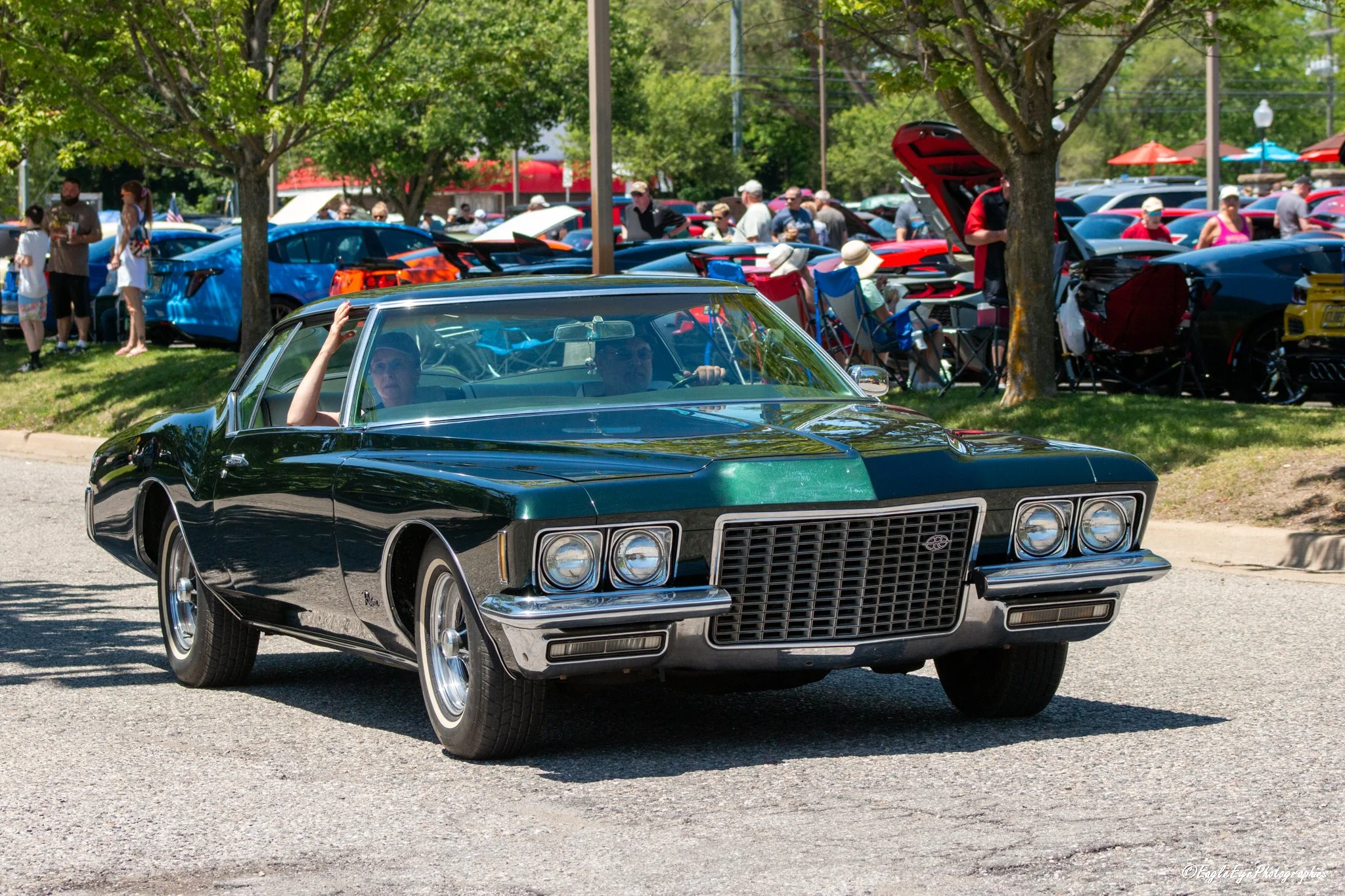 A vintage black car driving on a street during a parade or car show, with people and cars visible in the background, some under trees and umbrellas.