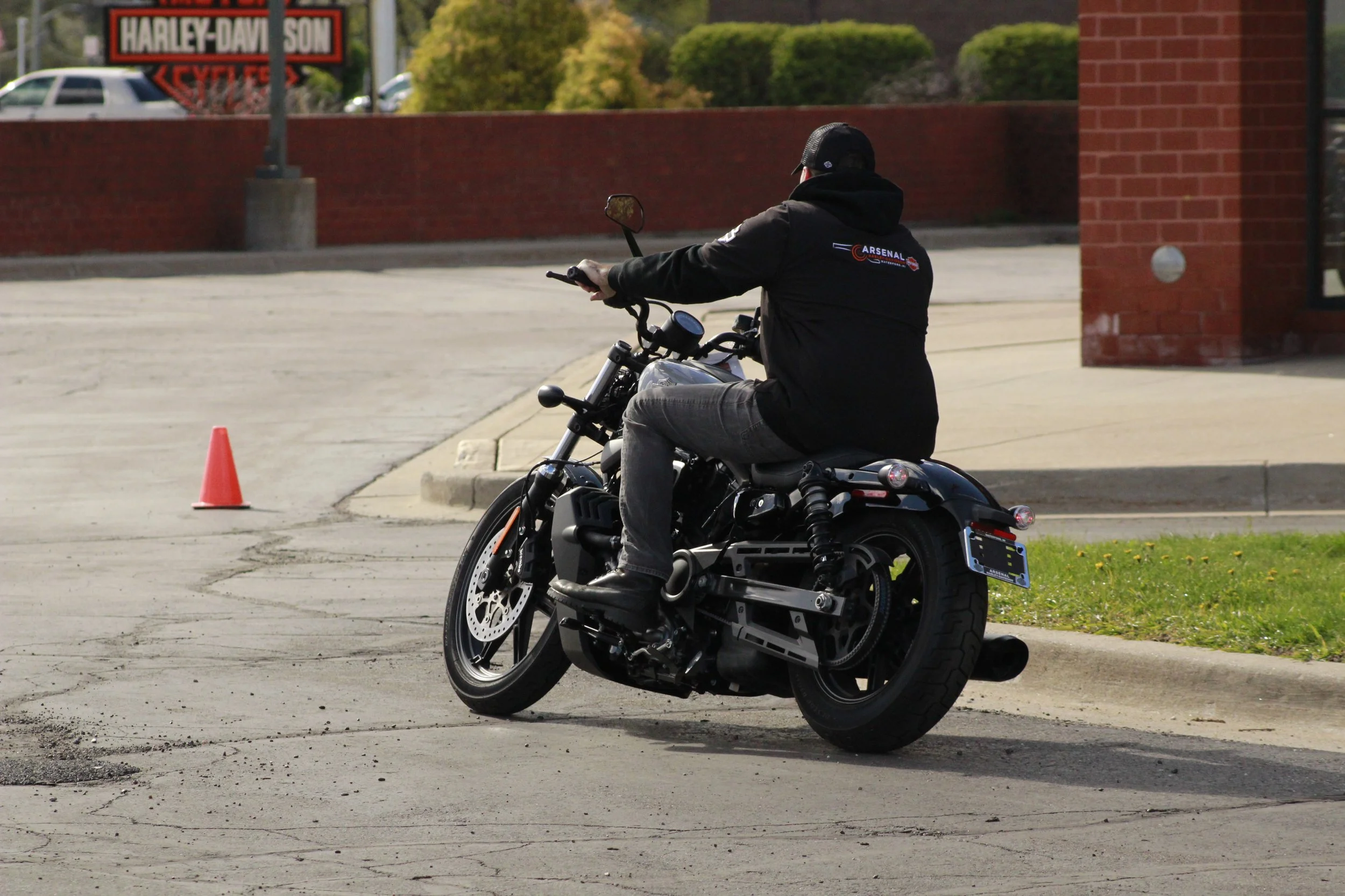 A person riding a black motorcycle on an urban street, wearing a black hoodie with a logo on the back, jeans, and a helmet, with a traffic cone nearby and a red brick building and a Harley-Davidson sign in the background.