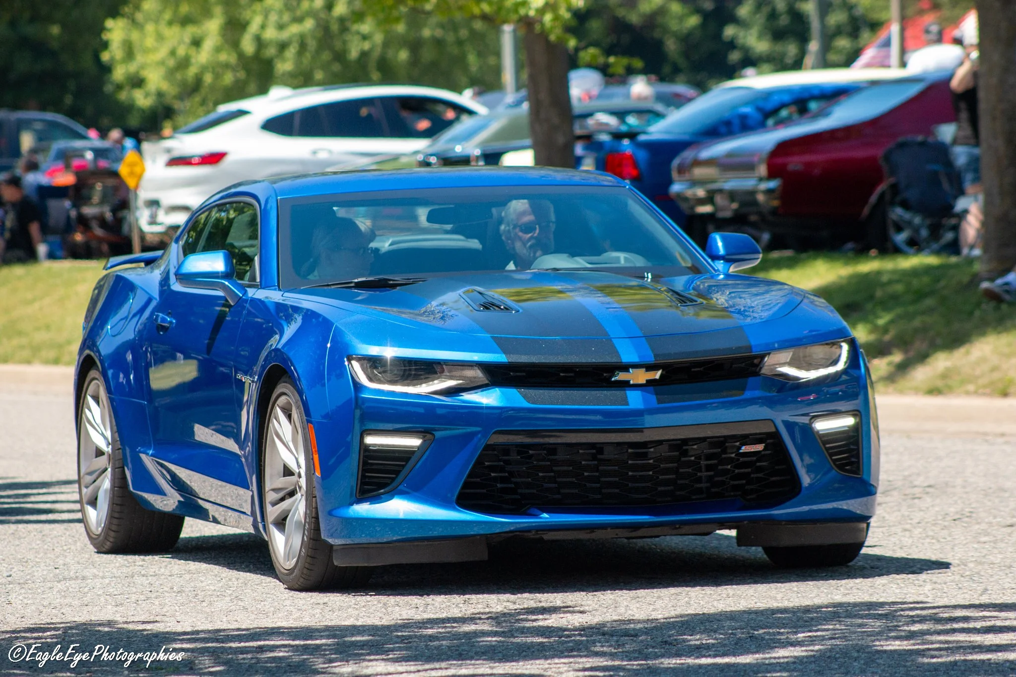 A blue Chevrolet Camaro SS sports car with black racing stripes on the hood, driving on a road during a car show.