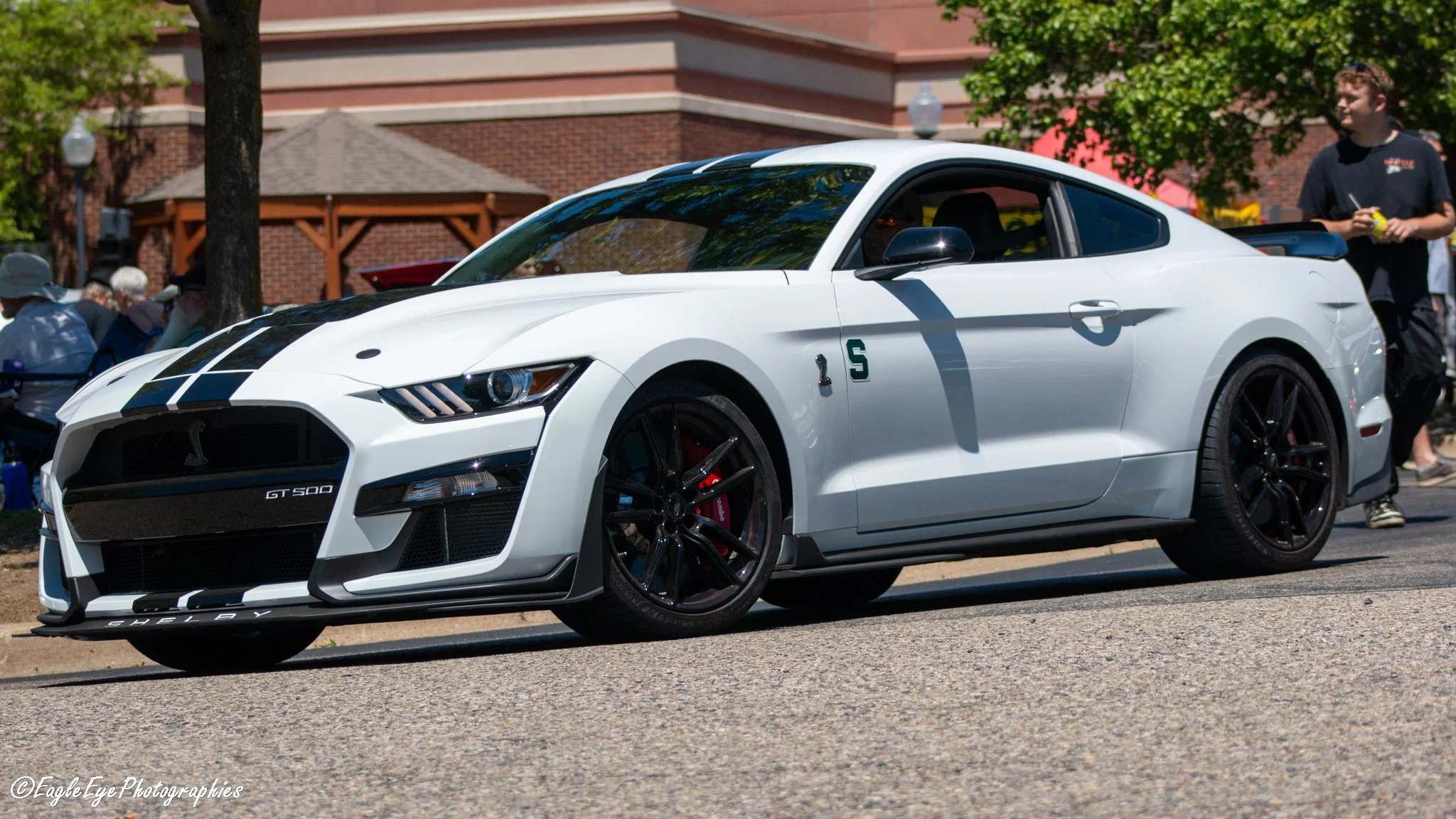 A white Shelby GT500 sports car with black racing stripes parked outdoors at a car show, with people and trees in the background.