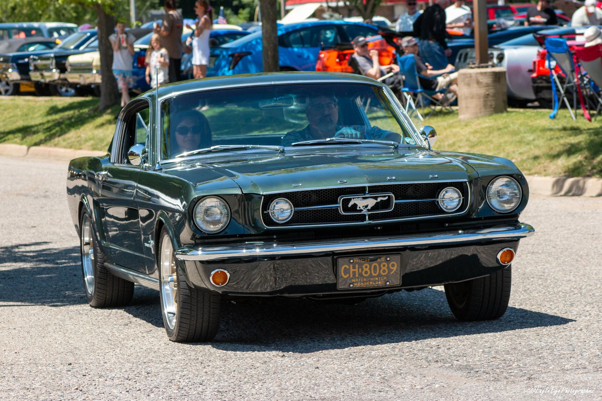A black classic Ford Mustang driving on a street during a car show event, with people in the background observing the lineup of parked cars.