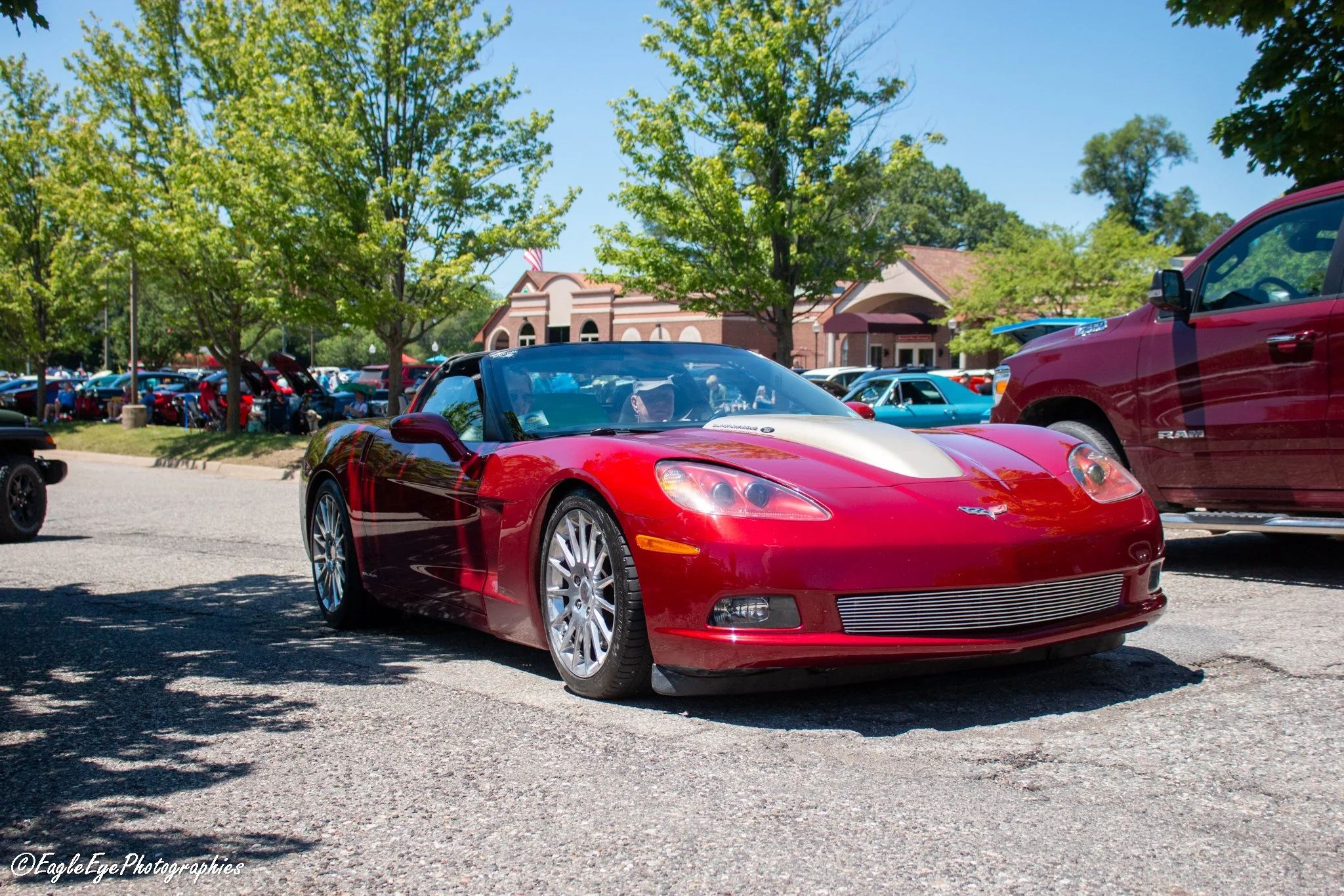 A red Chevrolet Corvette sports car parked in a lot with trees and other cars, a building in the background, and clear blue skies.