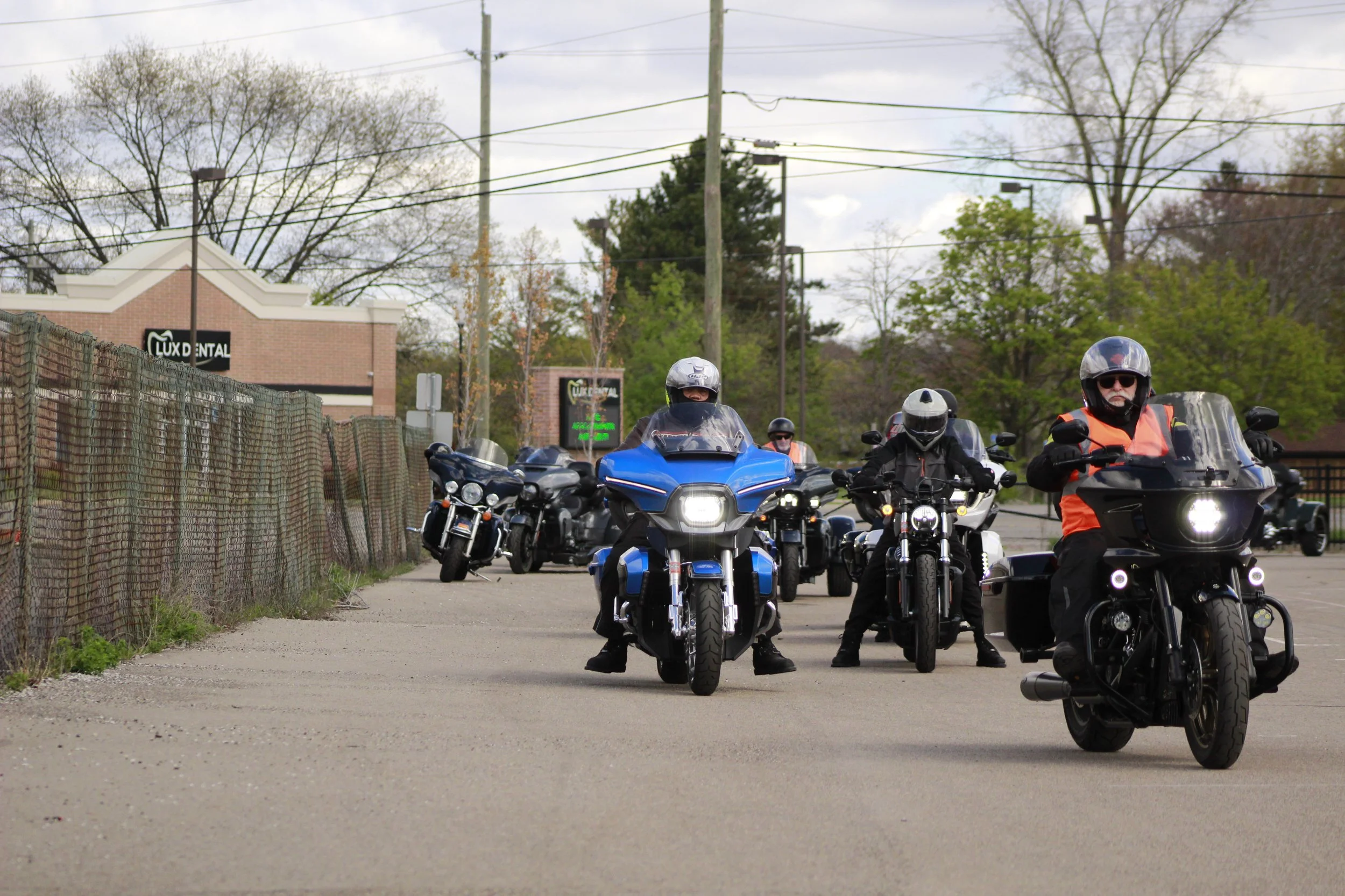 A group of motorcyclists riding on a road, wearing helmets and protective gear, with a building and trees in the background.