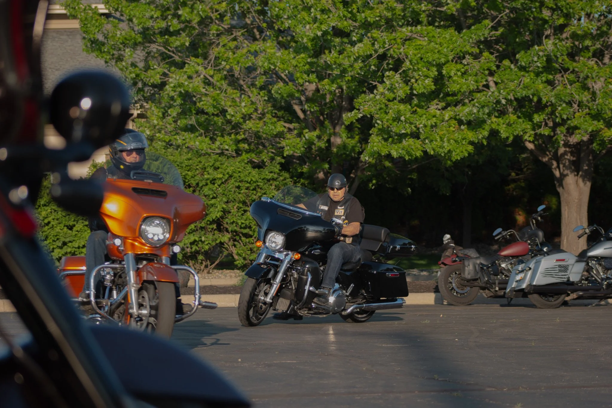 Motorcycles parked in a lot with two riders wearing helmets, one in black and the other in brown, during the day with green trees in the background.