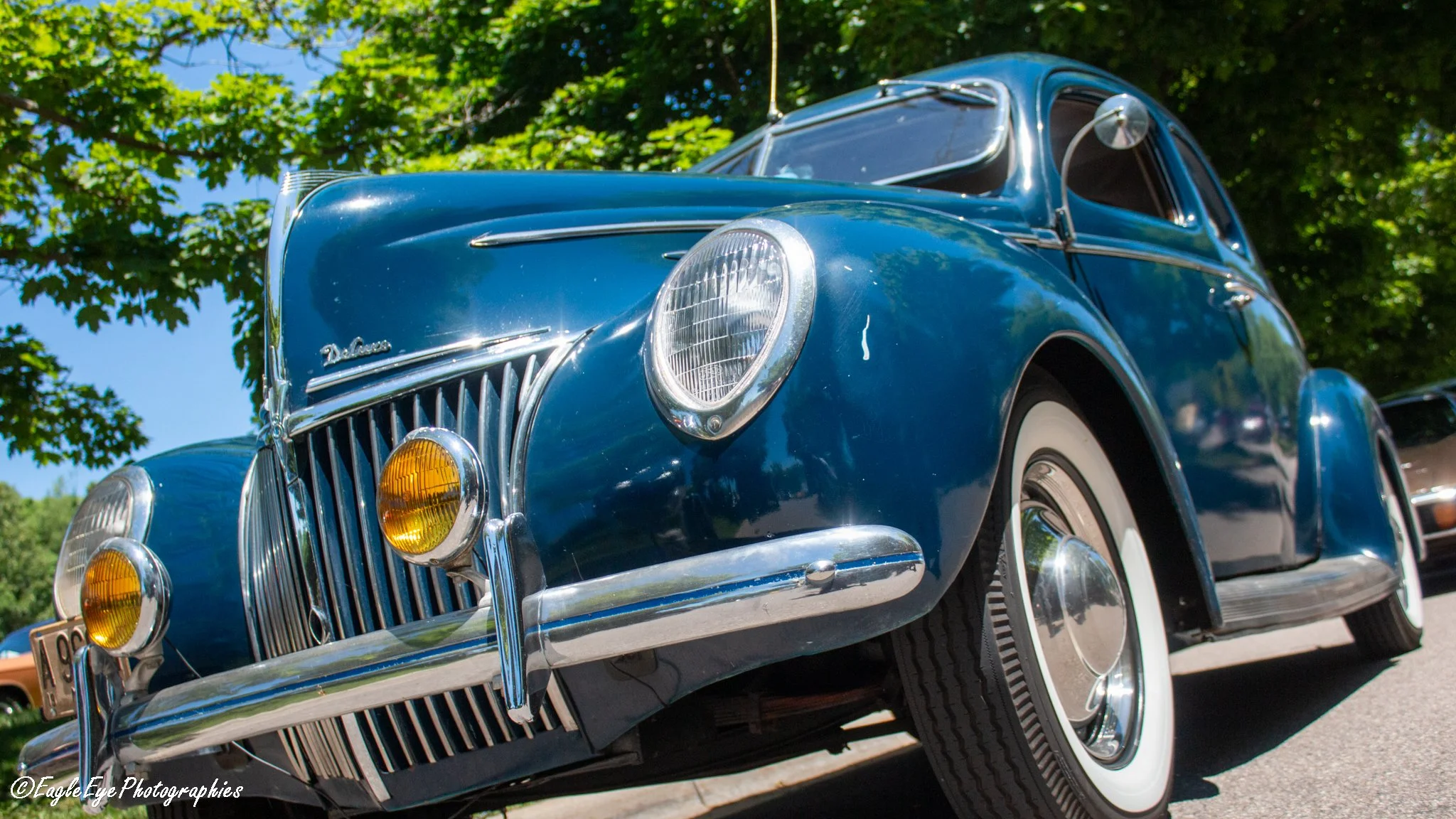 A vintage blue car with chrome accents and whitewall tires is parked outdoors under a tree with green leaves, with a clear blue sky in the background.