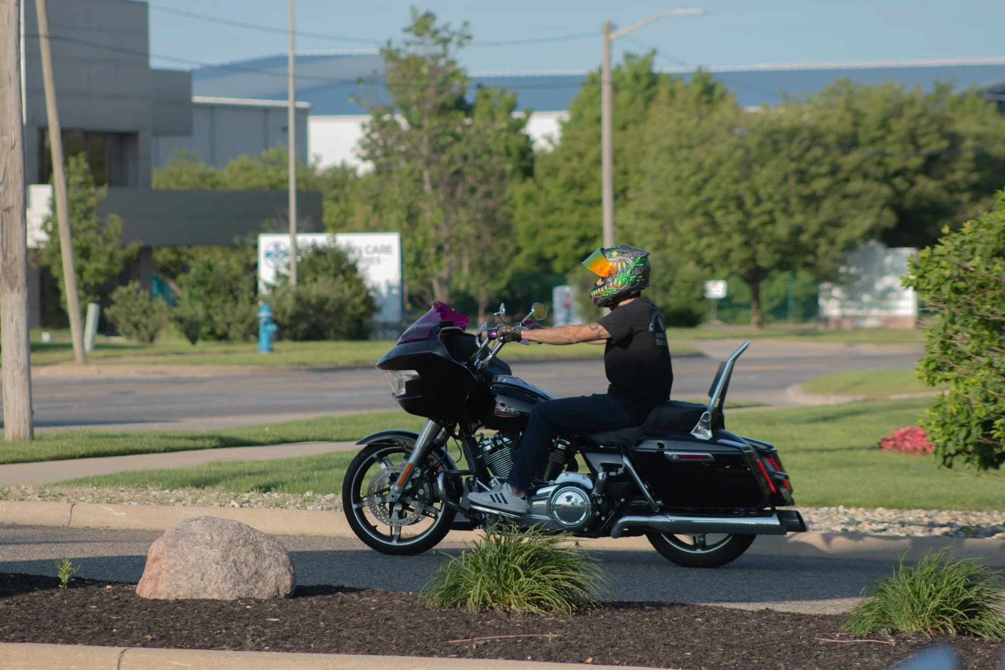 A person wearing a colorful helmet riding a black motorcycle on a street with greenery and buildings in the background.