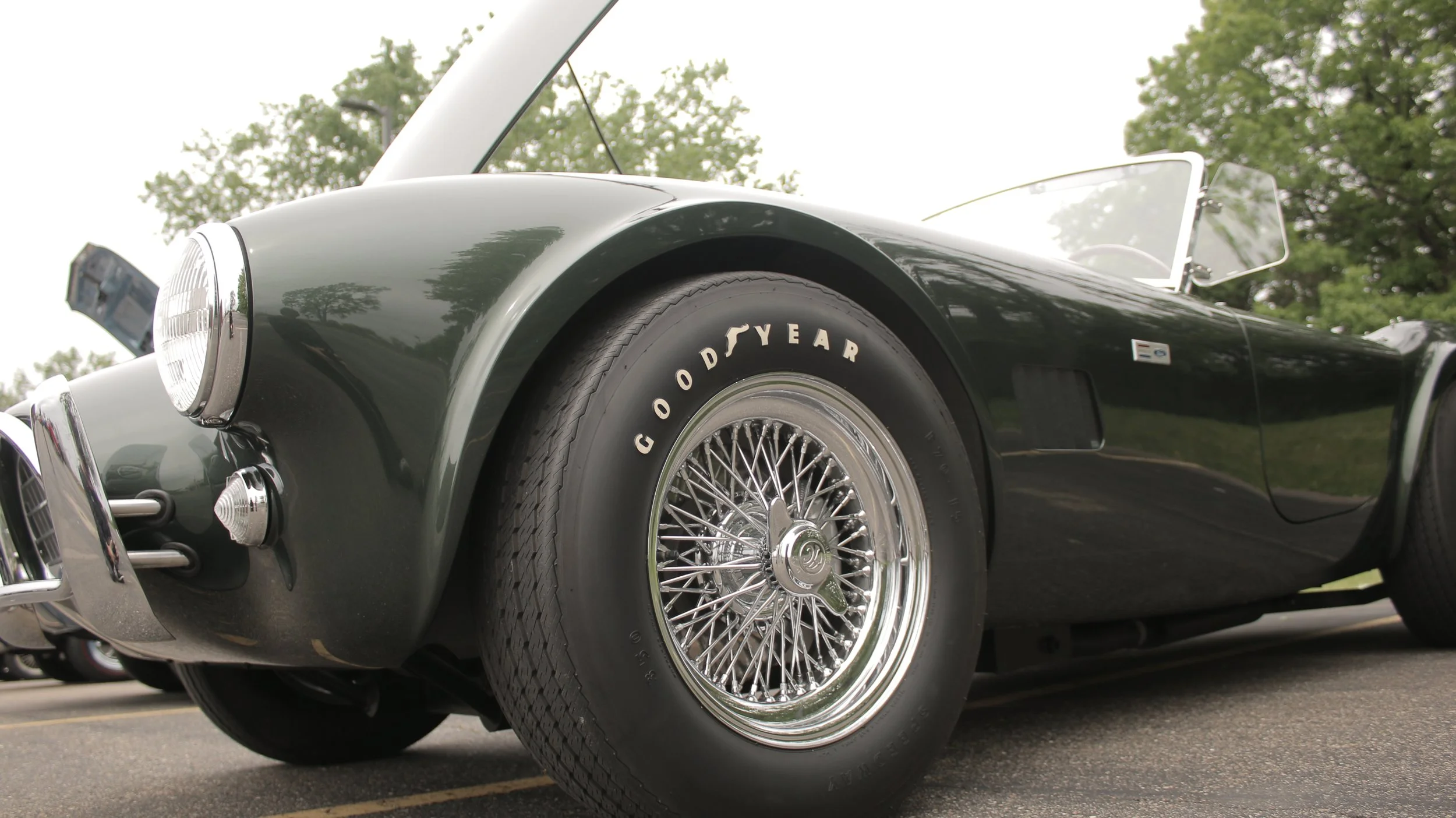 Close-up of a black vintage car with wire-spoke wheels and a Goodyear tire, parked on a street with green trees in the background.