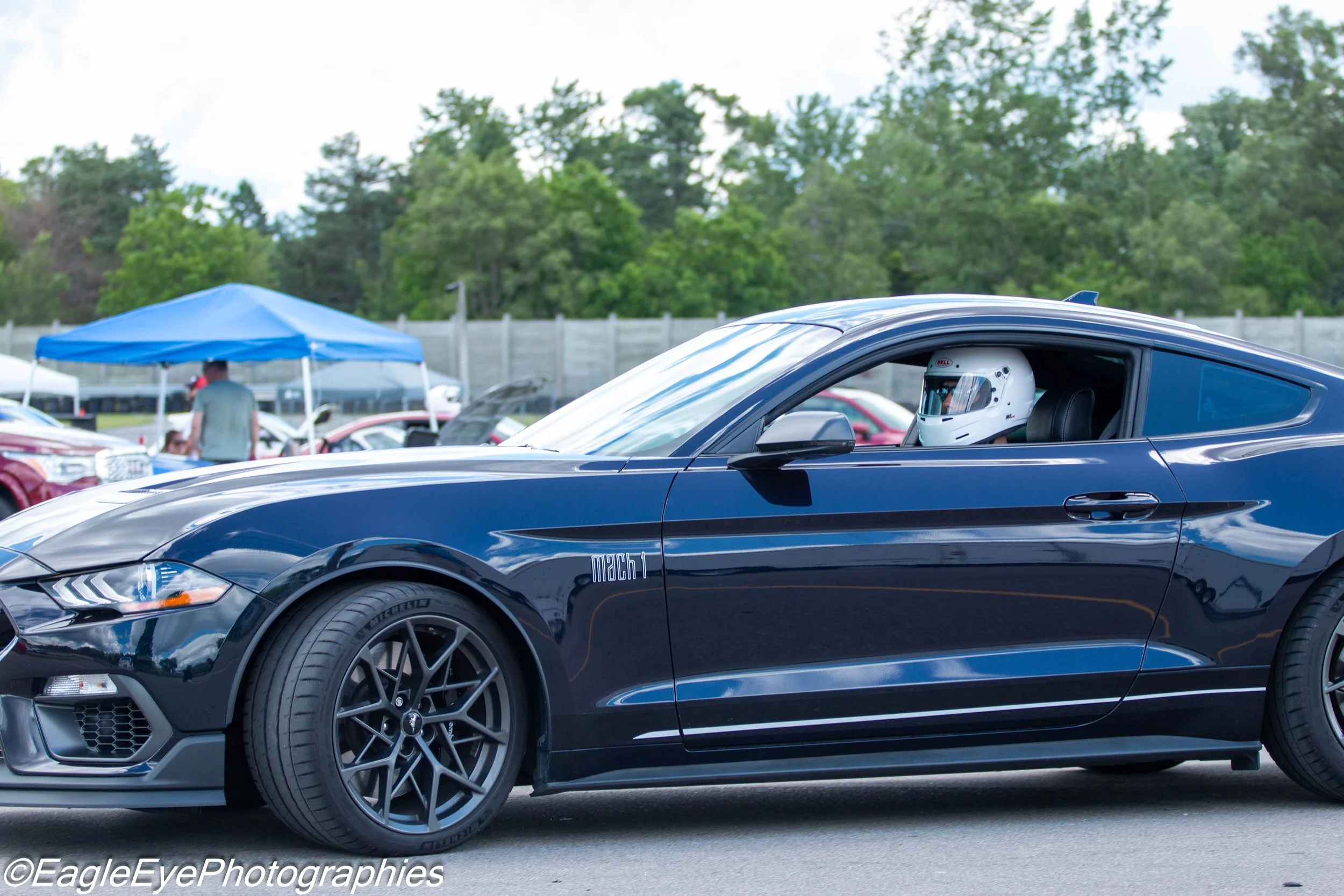 Close-up side view of a black Ford Mustang Mach 1 with a person wearing a white helmet inside, at an outdoor car show with other cars, a blue canopy, and trees in the background.