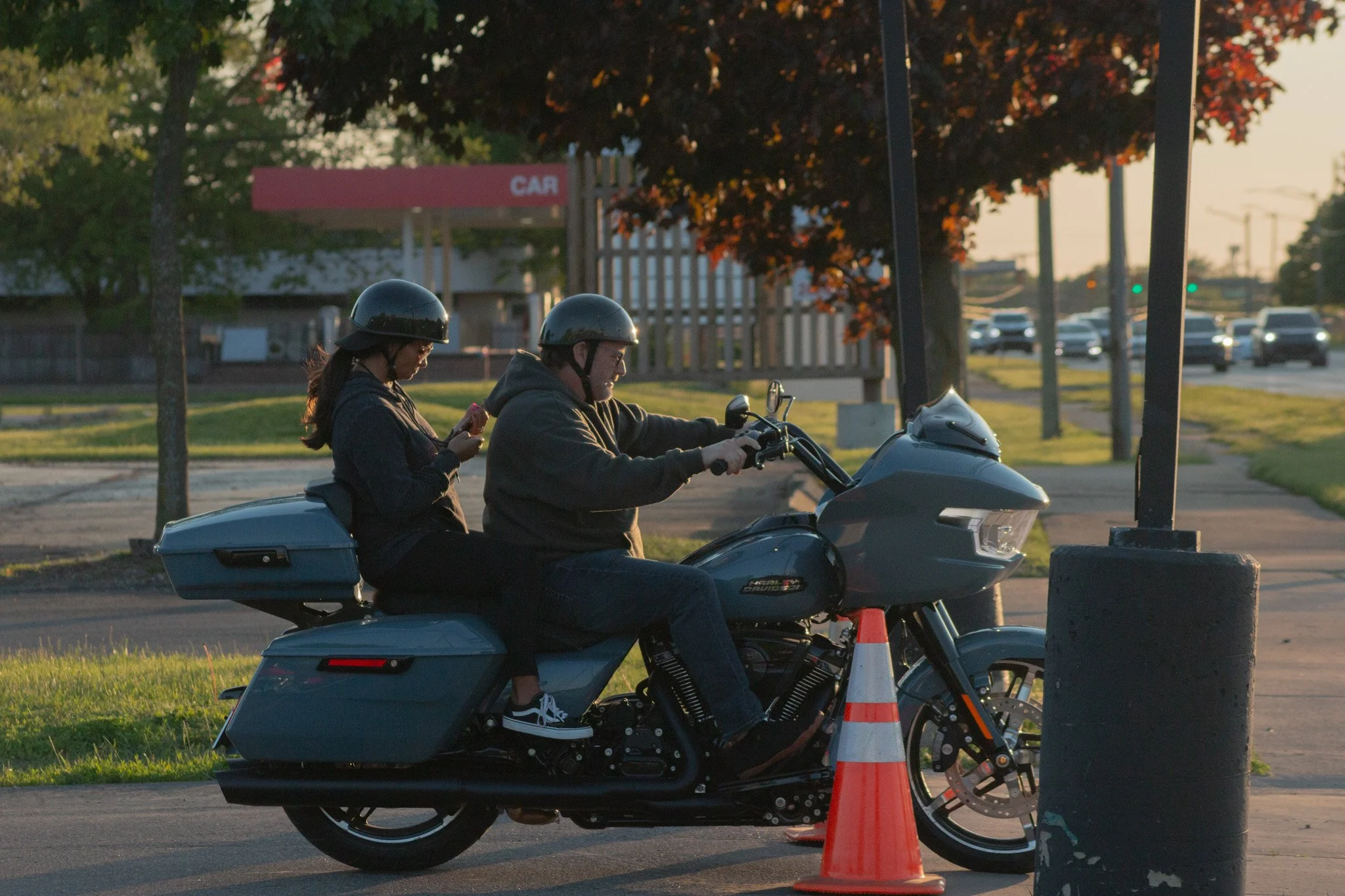 A man and woman on a motorcycle at a roadside checkpoint, with orange cones and a car parking sign in the background during sunset.