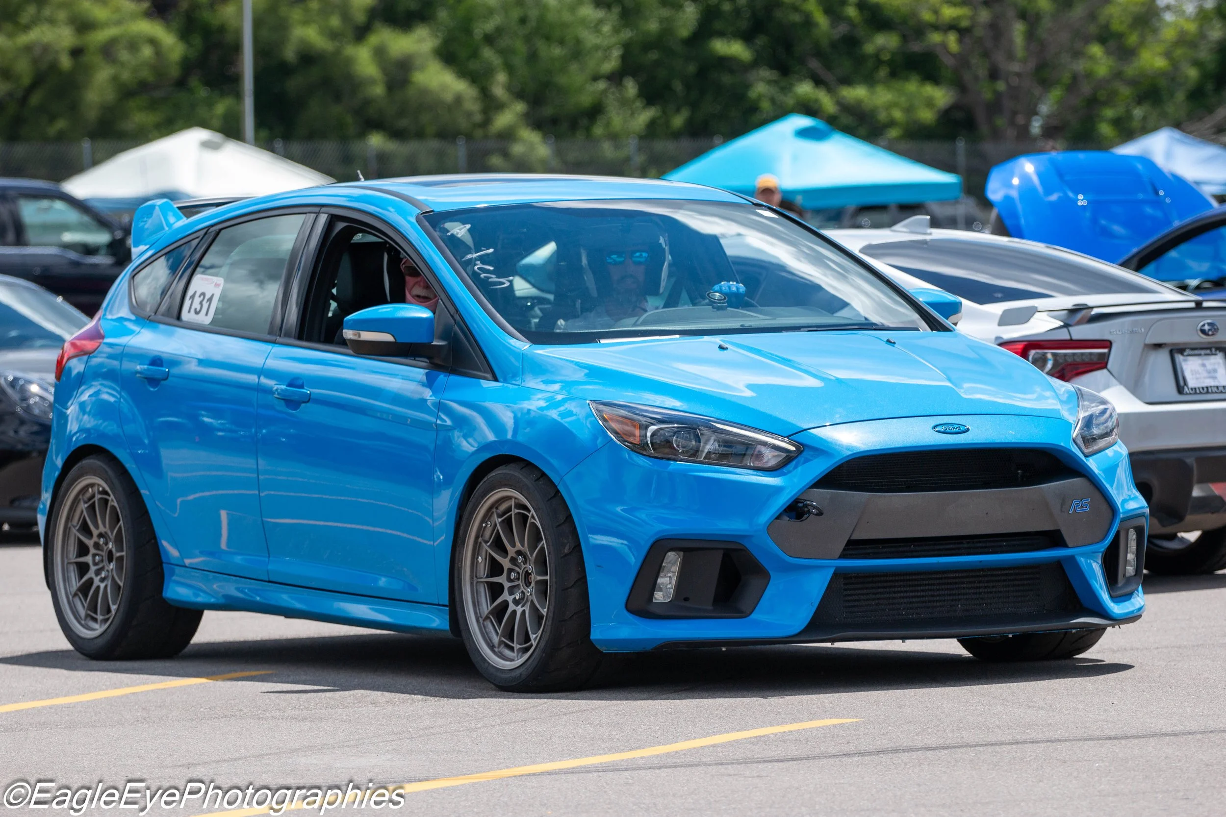 A bright blue Subaru car parked at a racing or car show event, with a person inside wearing glasses. Other vehicles are visible in the background, along with tents and trees, indicating an outdoor setting.