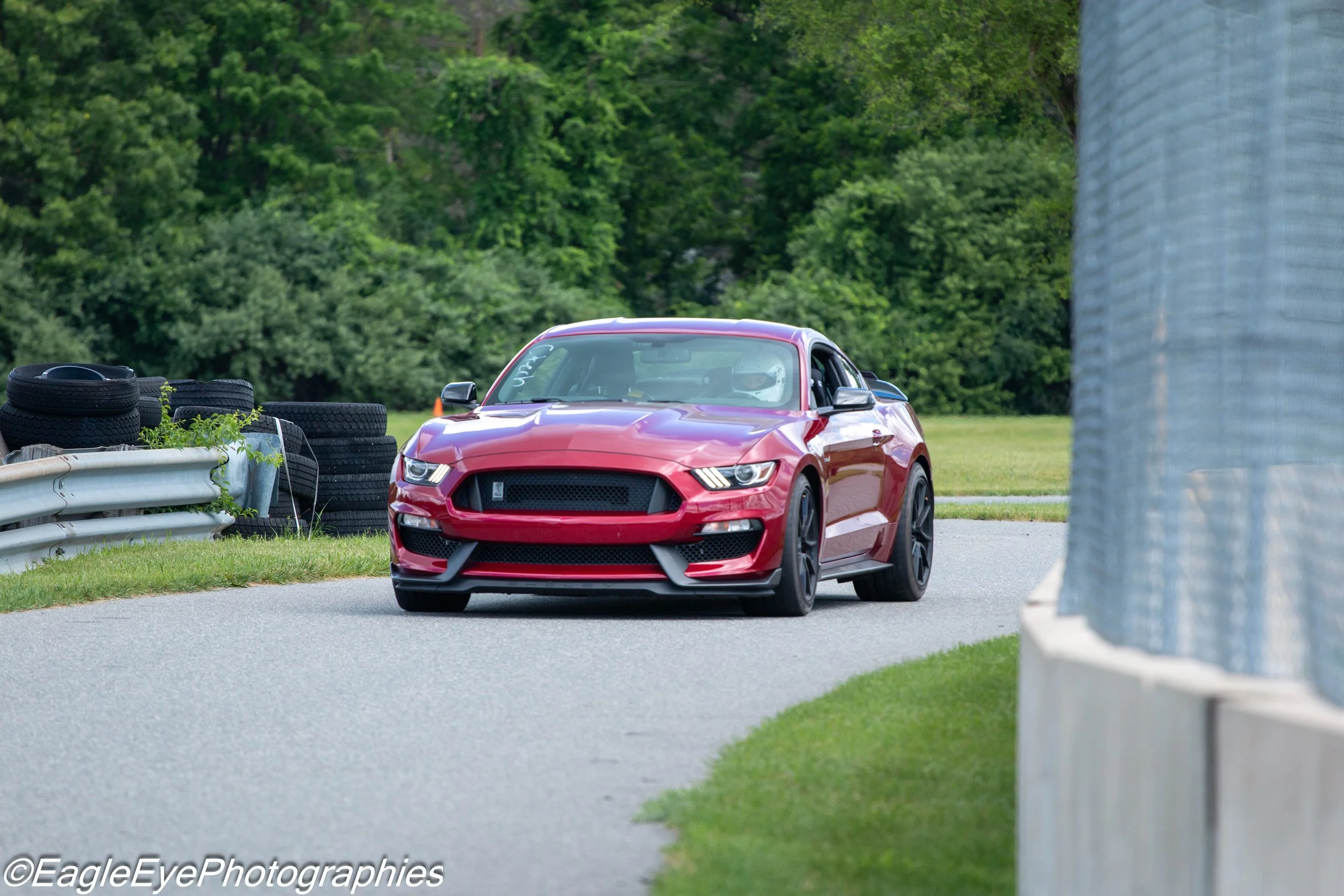 Red sports car driving past a row of tires and a concrete barrier on a road surrounded by green trees.