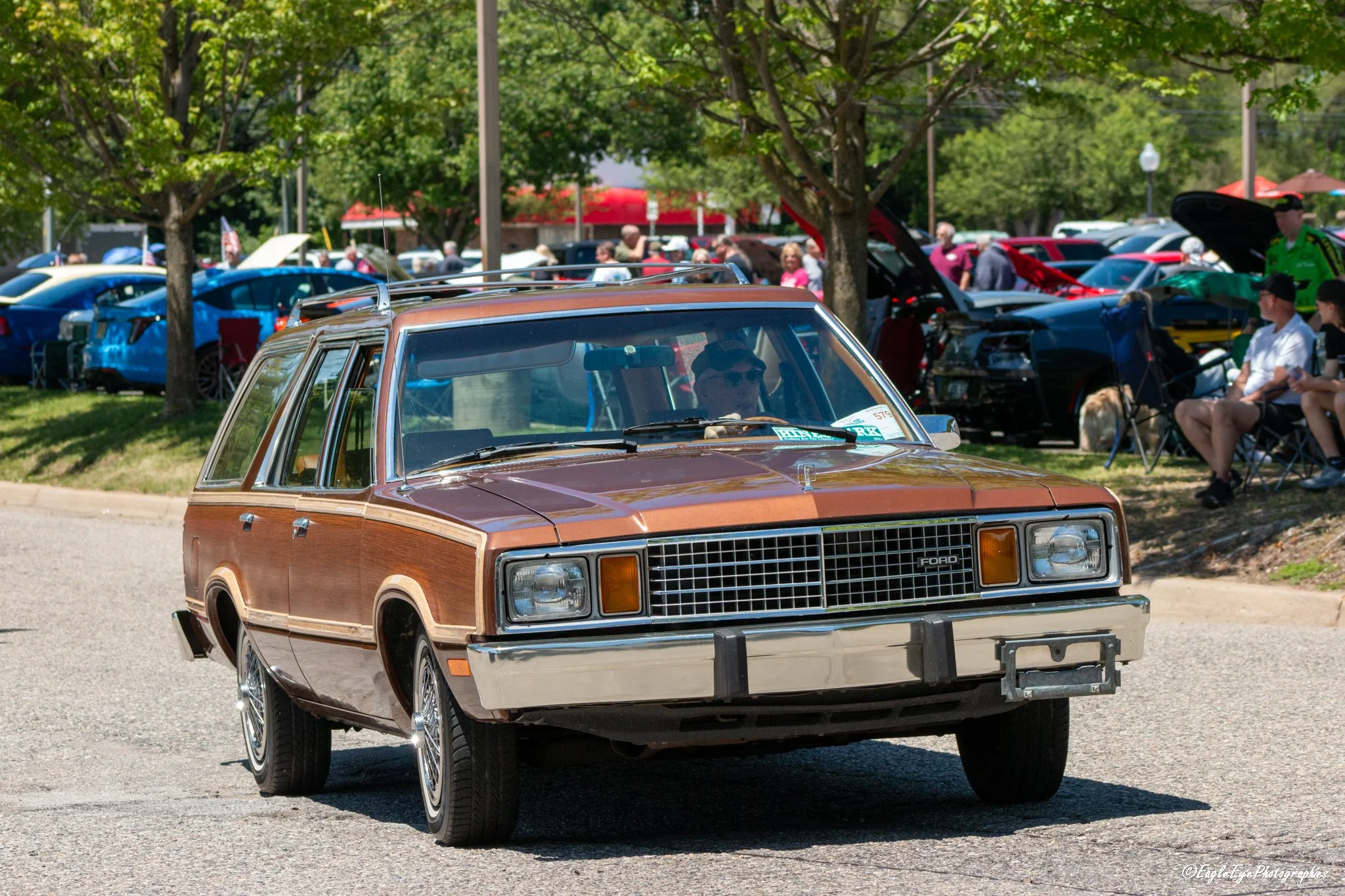 A vintage brown and tan Ford station wagon driving on a street during a classic car show, with a crowd of people and other cars displayed in the background.