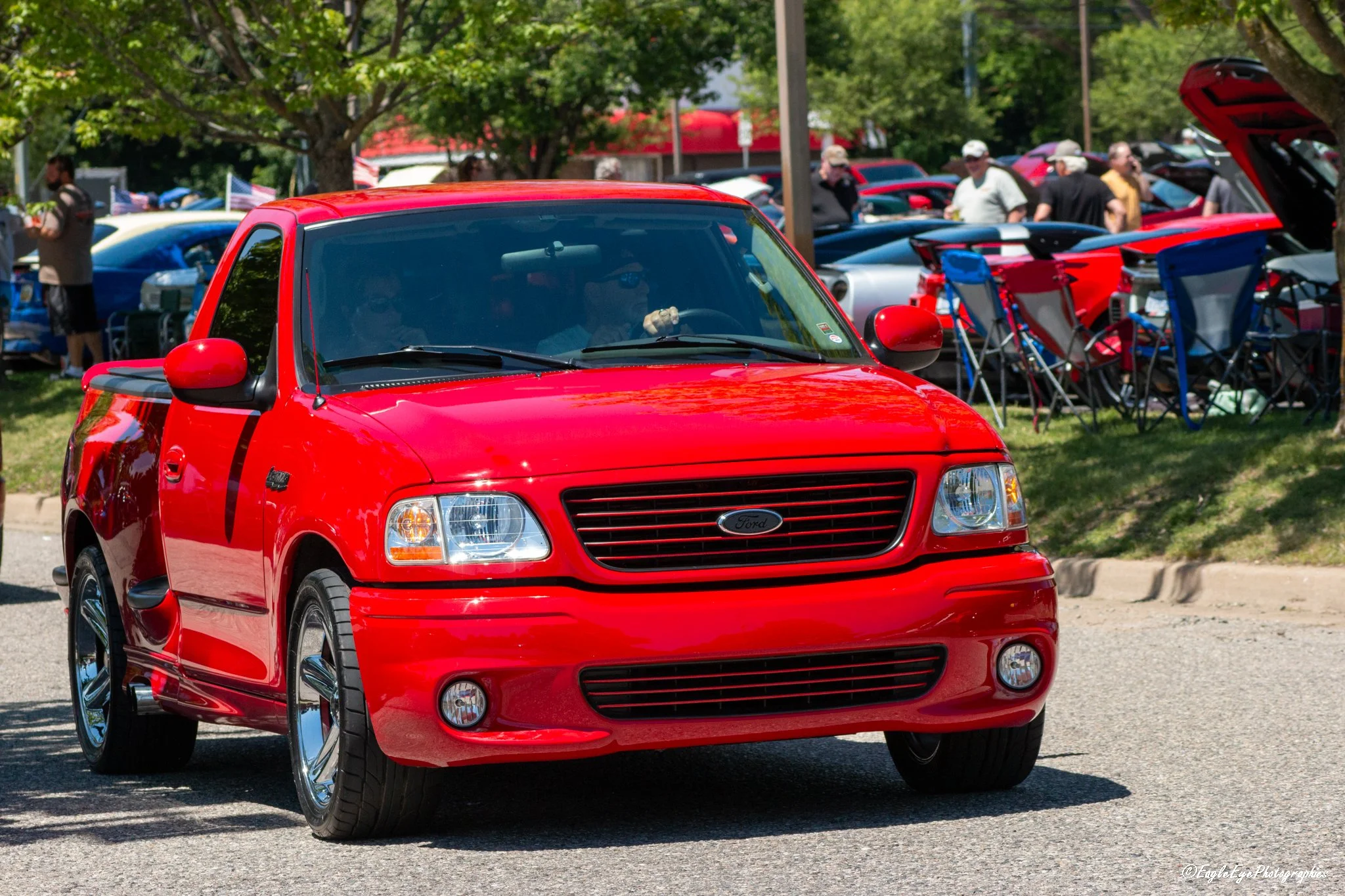 Red Ford pickup truck parked outdoors at a car show with people and other vehicles in the background.