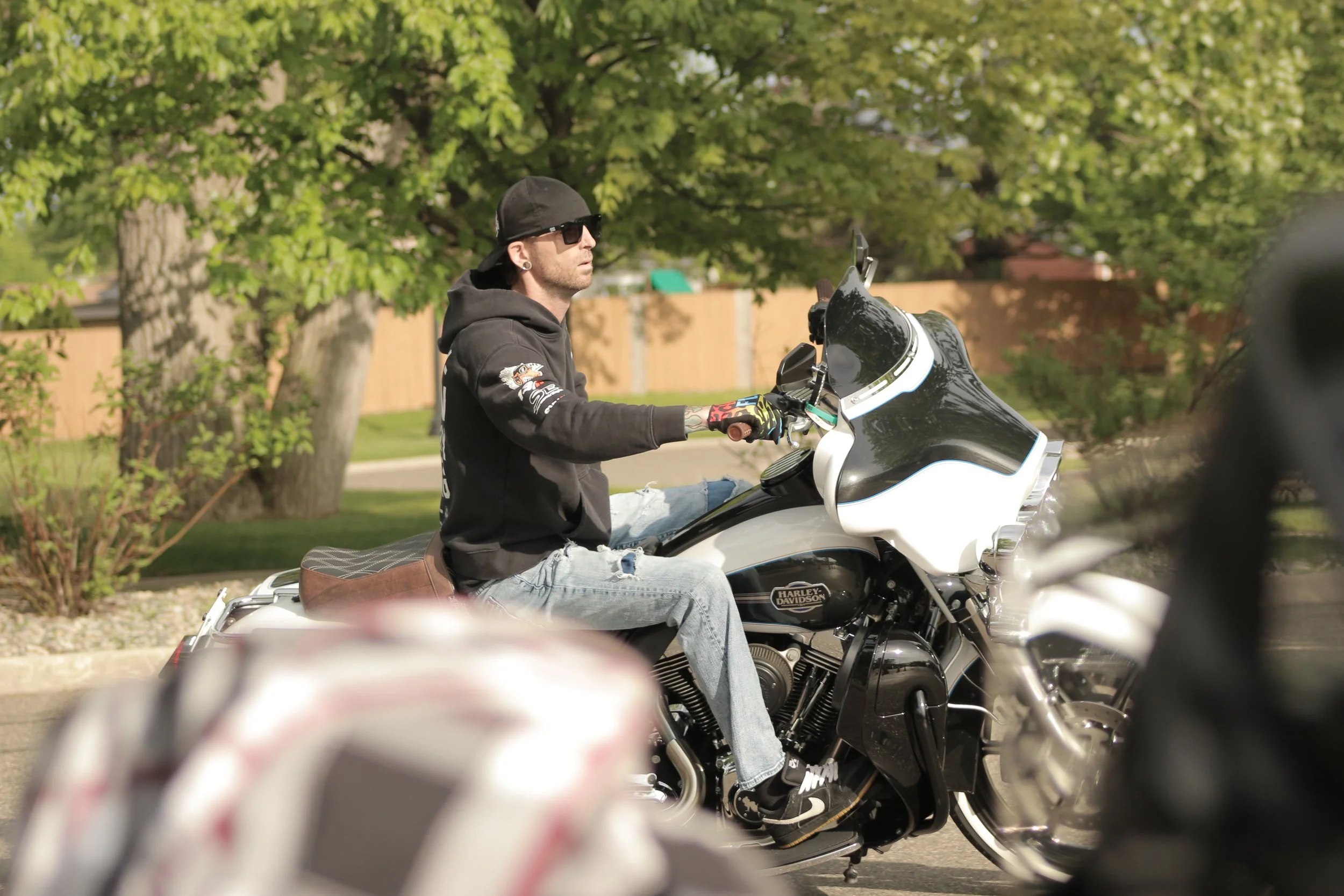 A man wearing sunglasses, a black hoodie, and a black cap is riding a black and white Harley Davidson motorcycle outdoors on a sunny day with green trees in the background.