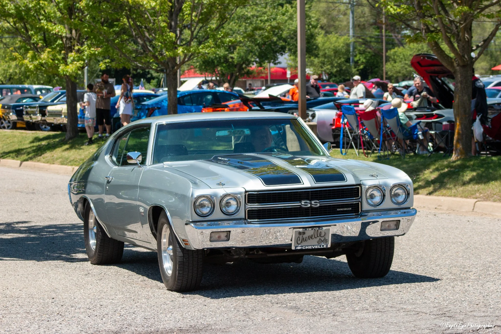 A classic silver Chevrolet Chevelle SS muscle car with black racing stripes parked at an outdoor car show with other cars and people in the background under trees.