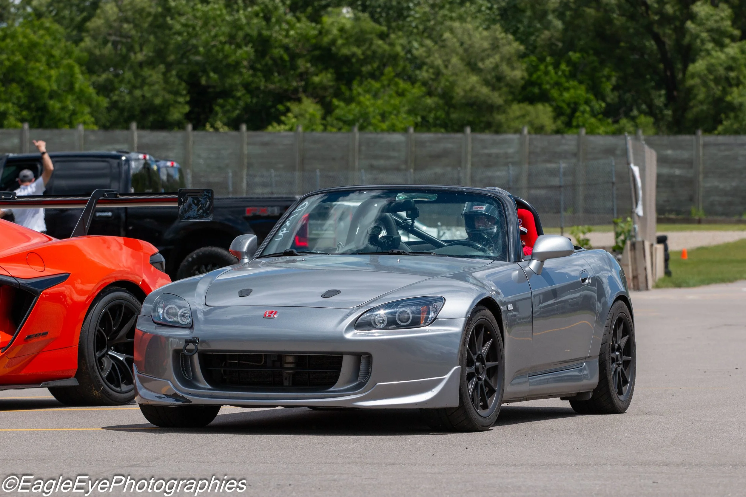 A silver Honda S2000 sports car parked among other vehicles at a racing event, with a person in racing gear visible inside and a person waving in the background near a black truck.