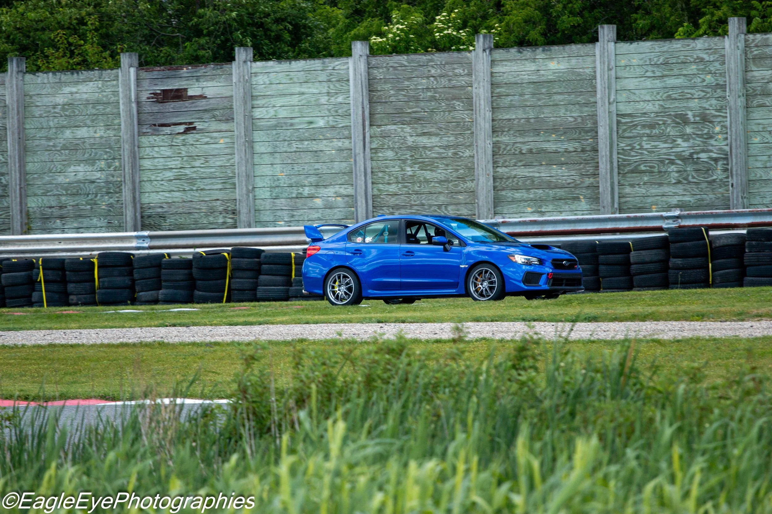 A blue sports car on a racing track with barriers, a tall wooden fence, and lush green trees in the background.