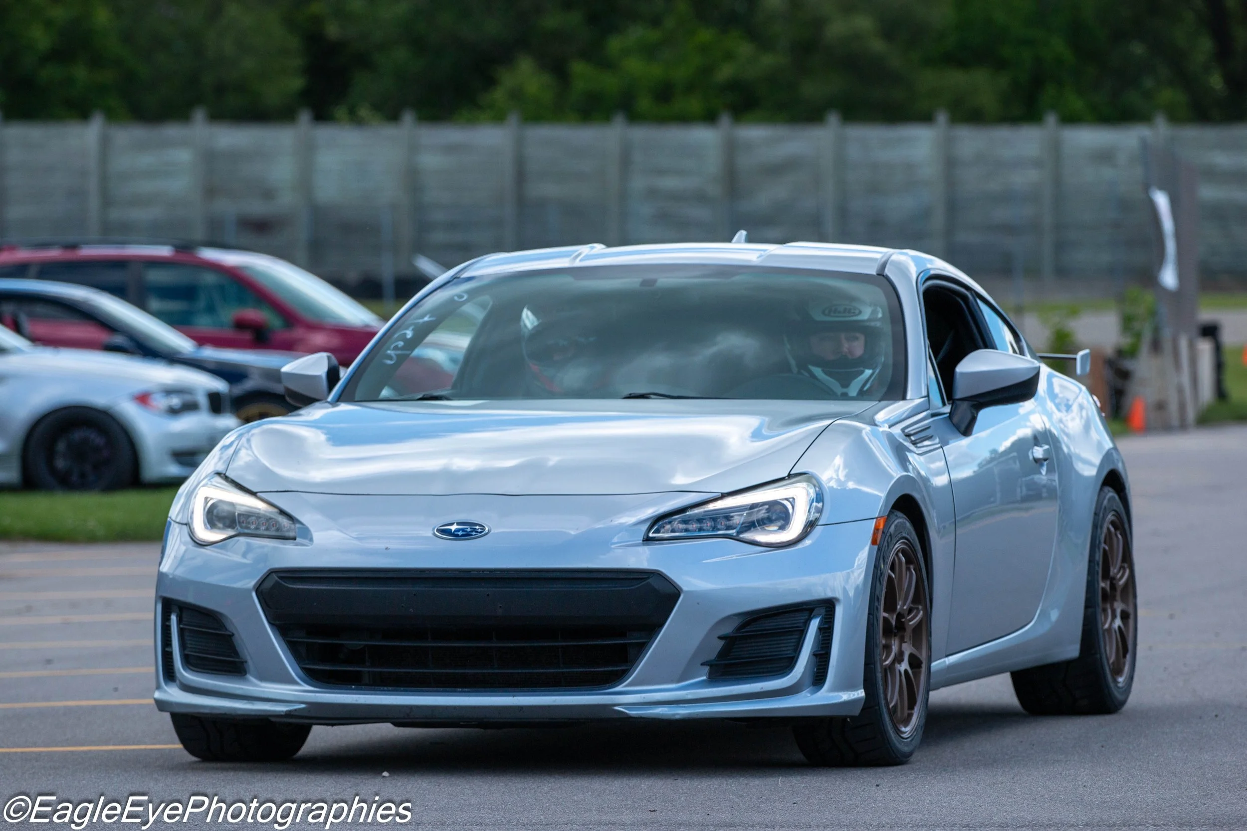 Gray sports car with a driver wearing a helmet in a parking lot, surrounded by other cars and a wooden fence in the background.