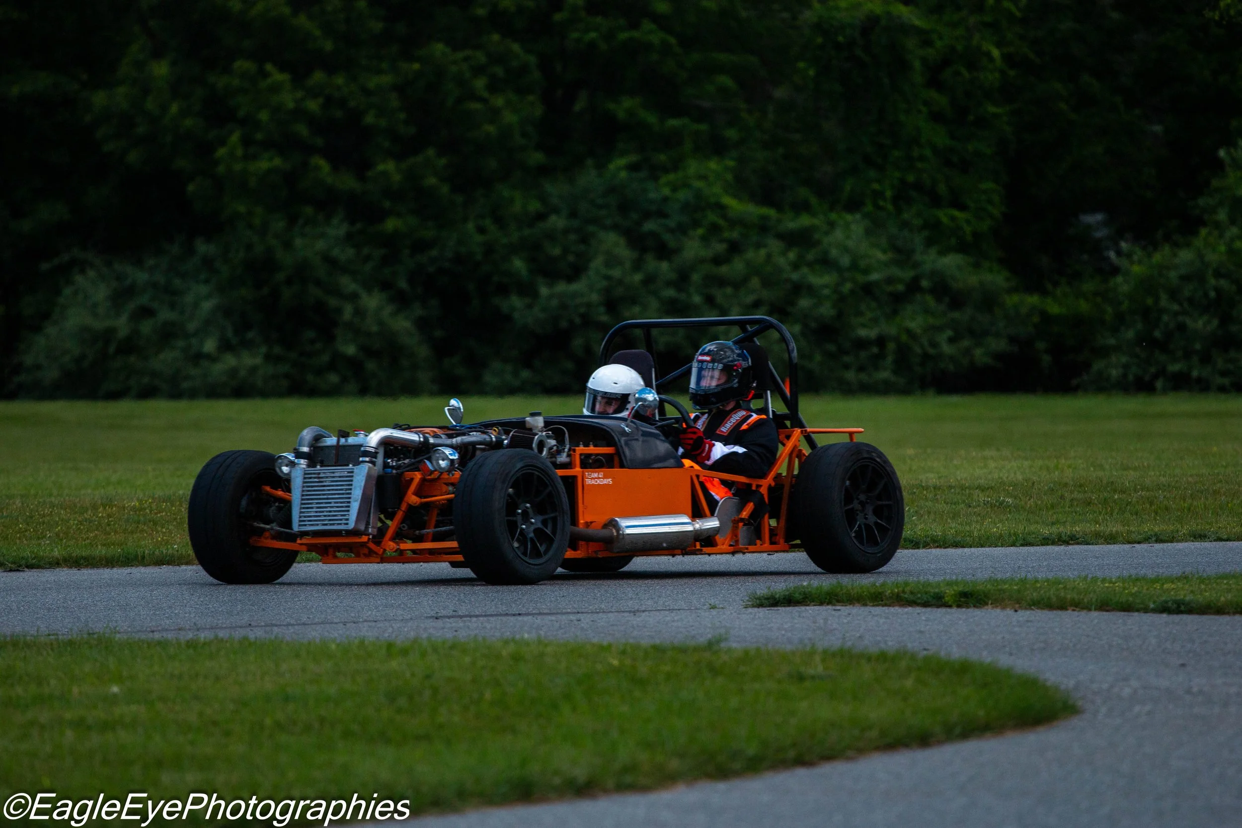 An orange racing go-kart on a track with two drivers wearing helmets and racing gear, surrounded by green trees.