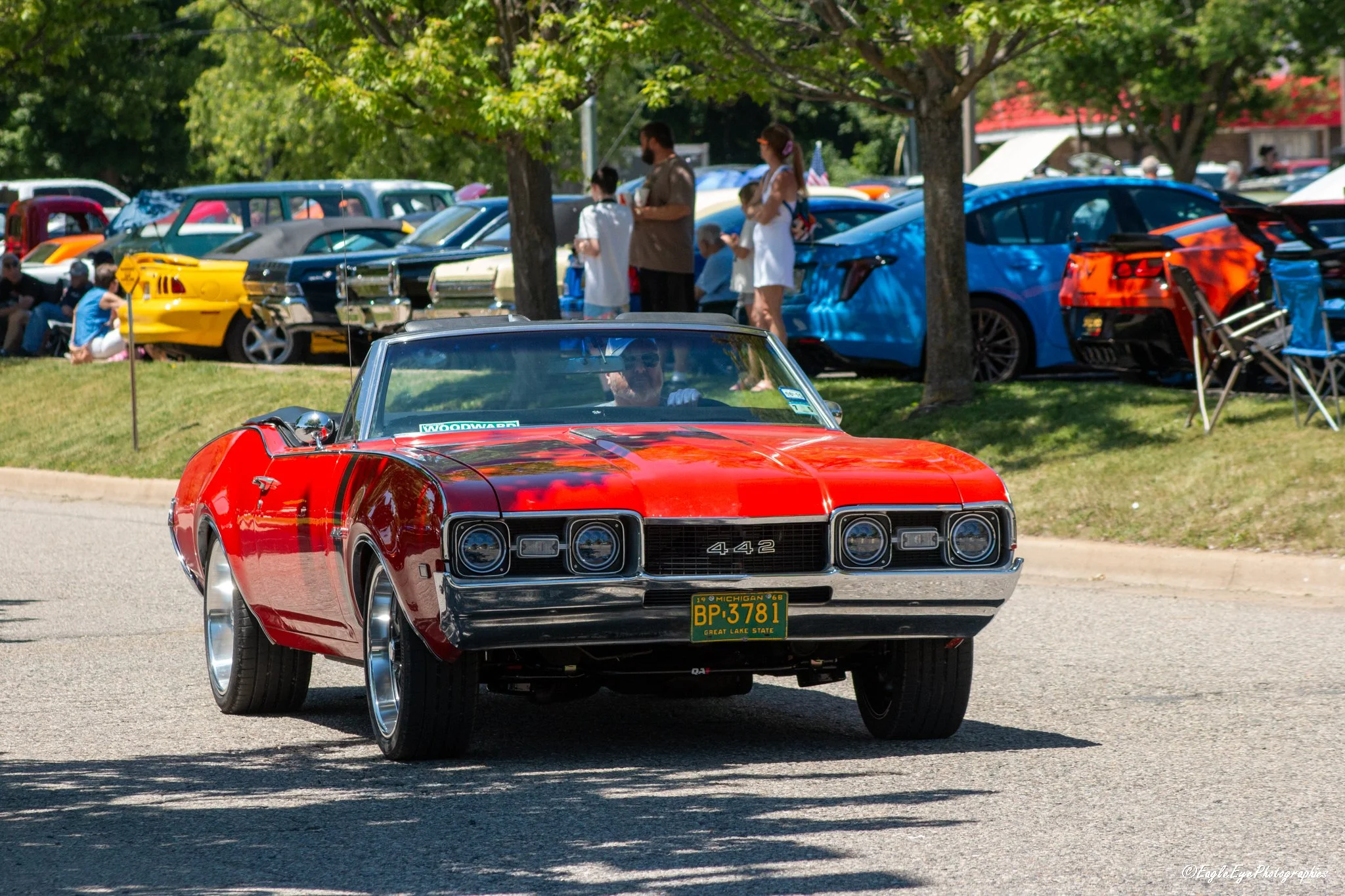 Red vintage Chevrolet Corvette with Michigan license plate BP 3781 driving past a car show with various classic and modern cars parked under trees.