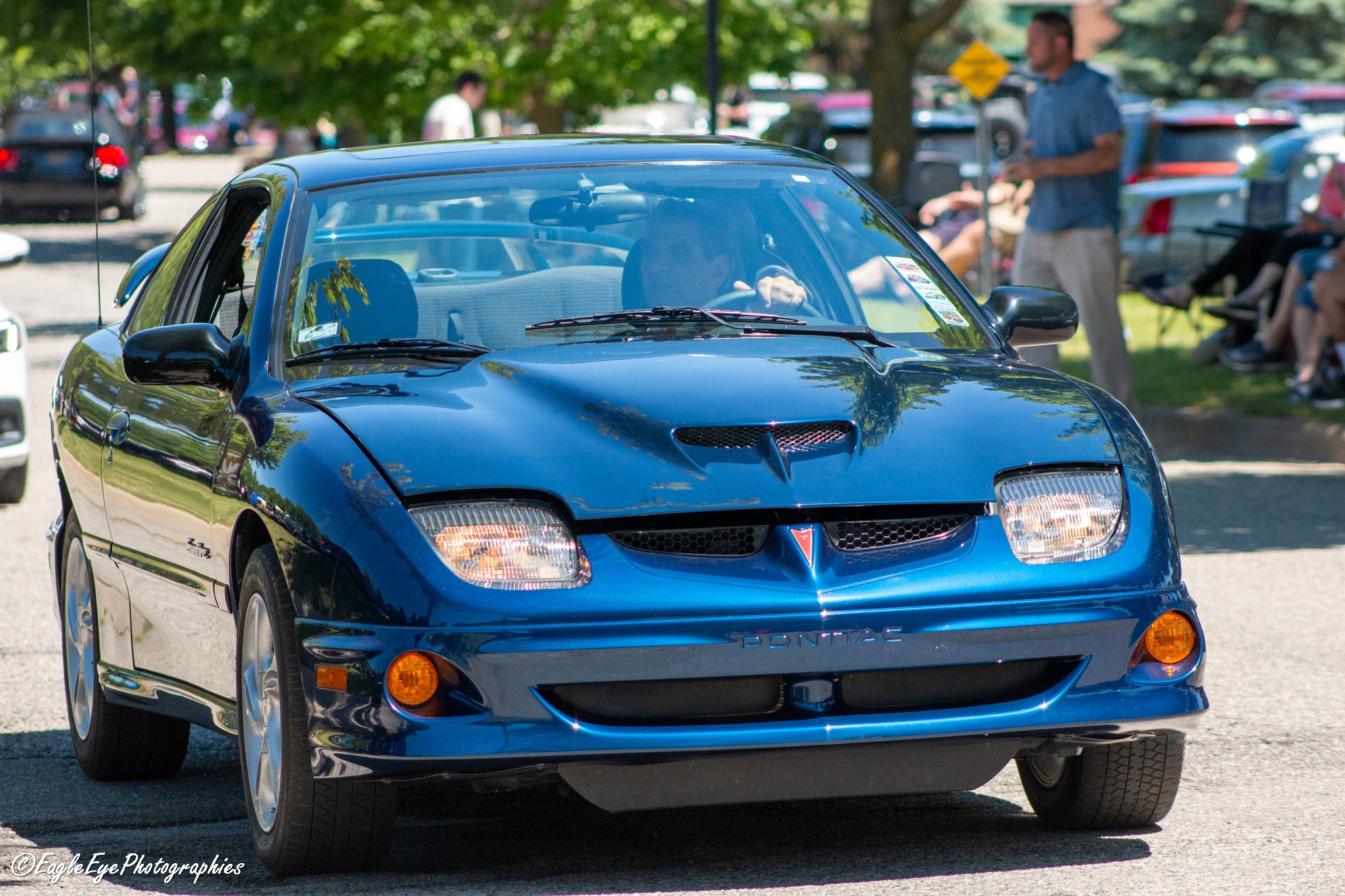 Front view of a shiny blue Pontiac firebird sports car parked outdoors with trees and people in the background.