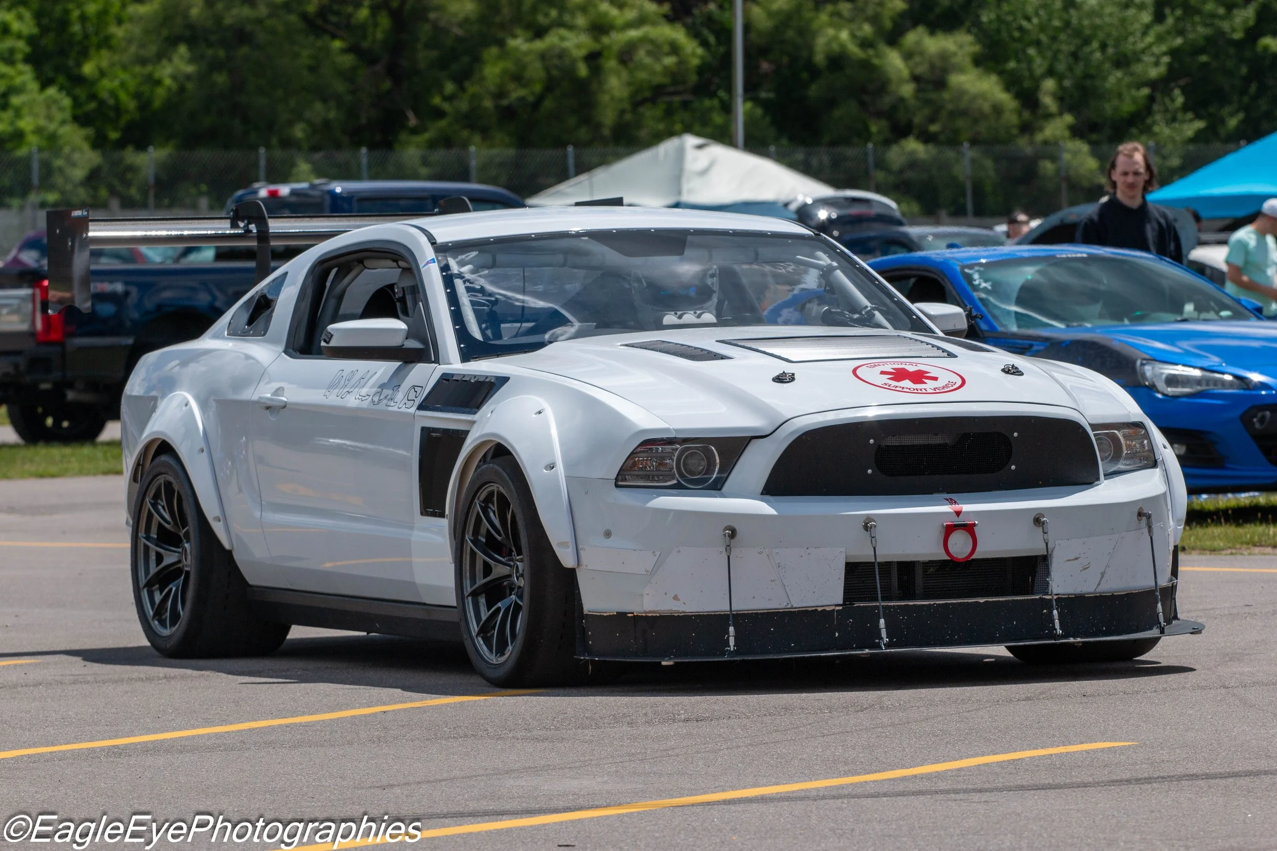 A white race car with a red support vehicle decal on the hood, parked in a lot during a car event. The car has a large rear wing and specialized wheels, with other cars and people visible in the background, under a cloudy sky.