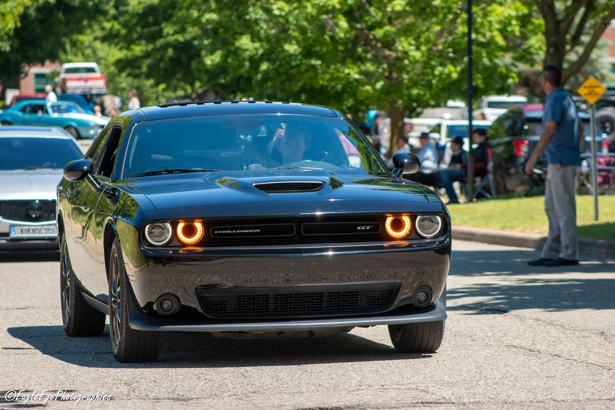 A black Dodge Challenger GT sports car parked on a street, with people and trees in the background during a car show or gathering.