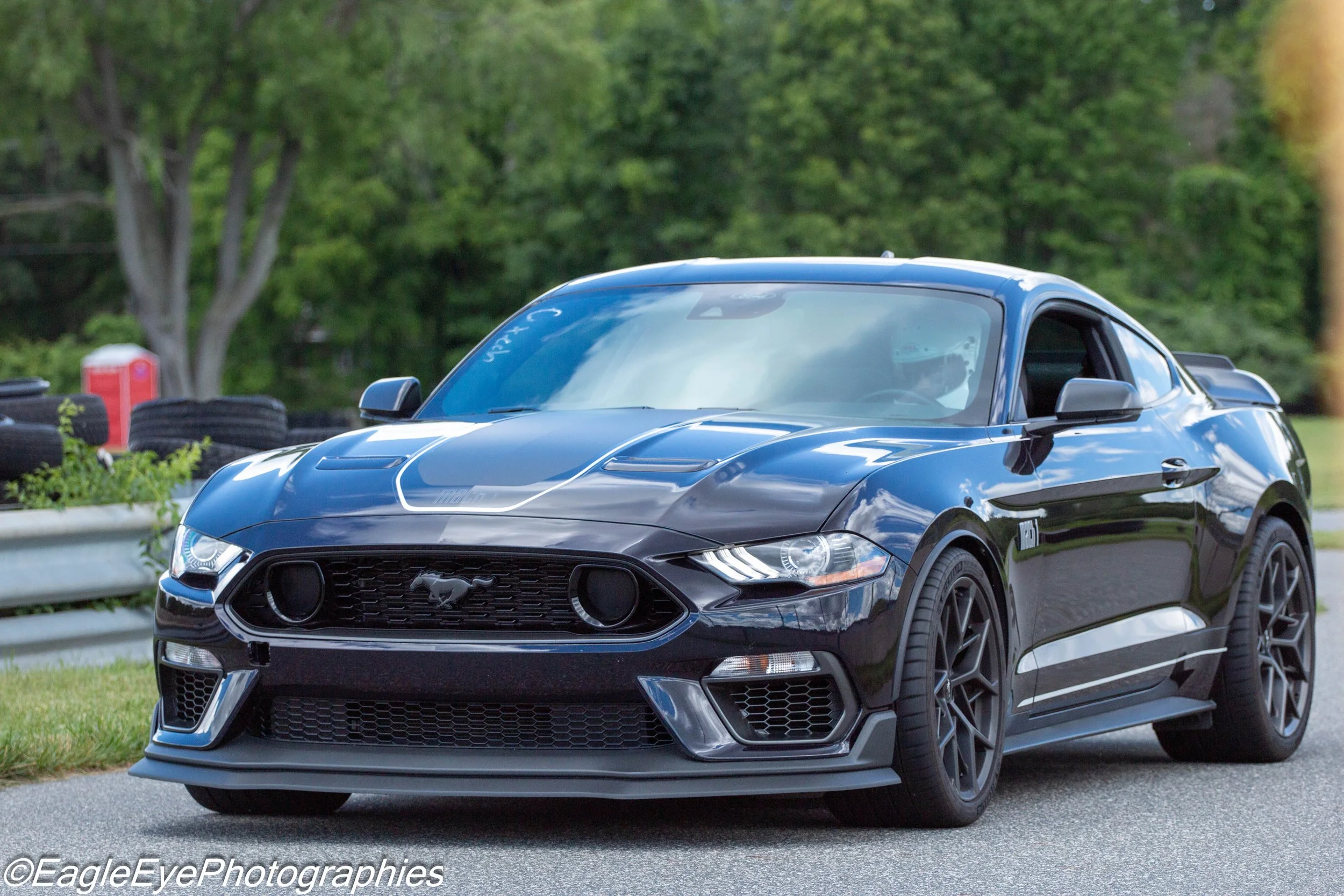 A black Ford Mustang GT sports car parked outdoors on a paved surface with green trees and tires stacked in the background.