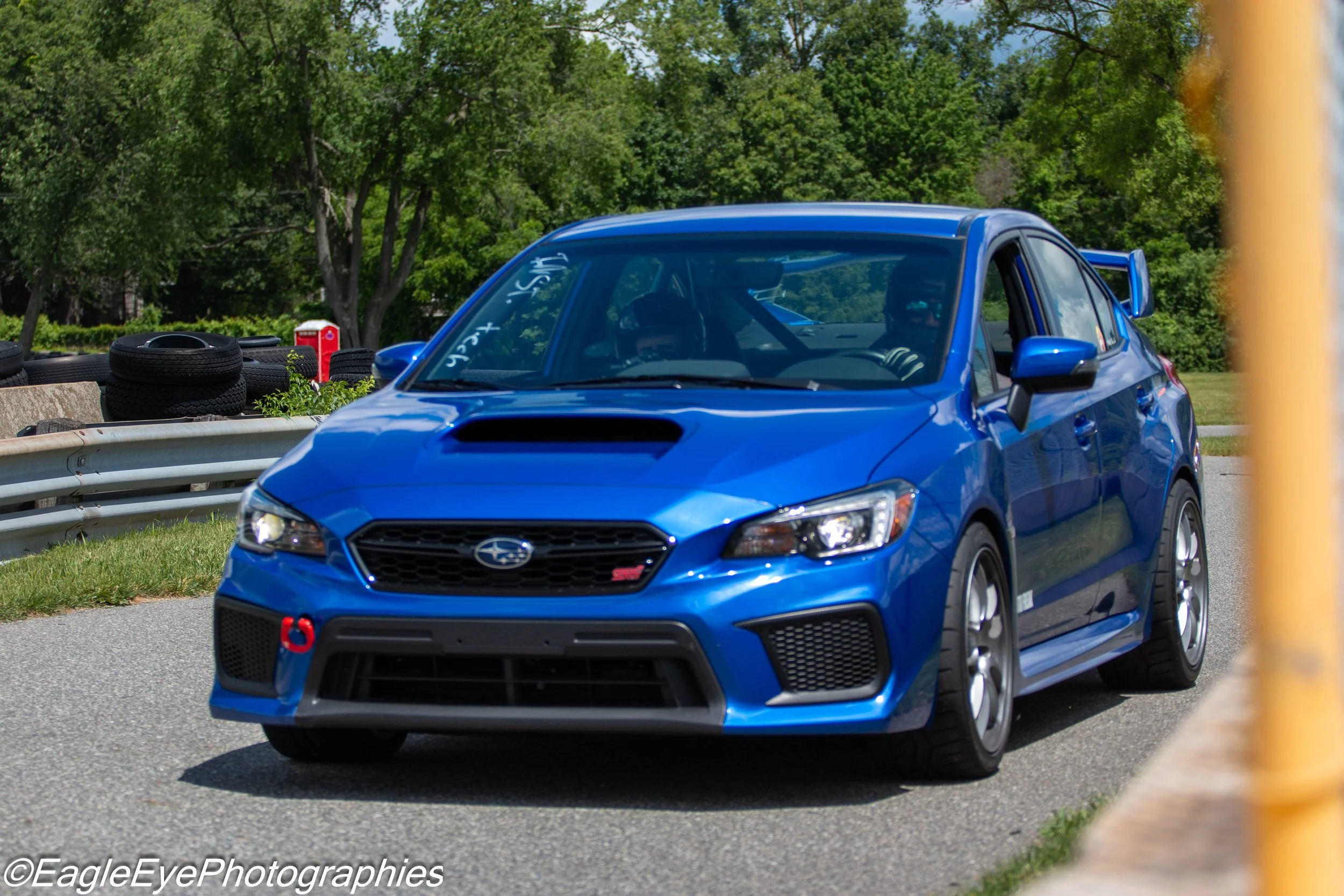 A blue Subaru WRX STI driving on a road, with a stack of tires and trees in the background.