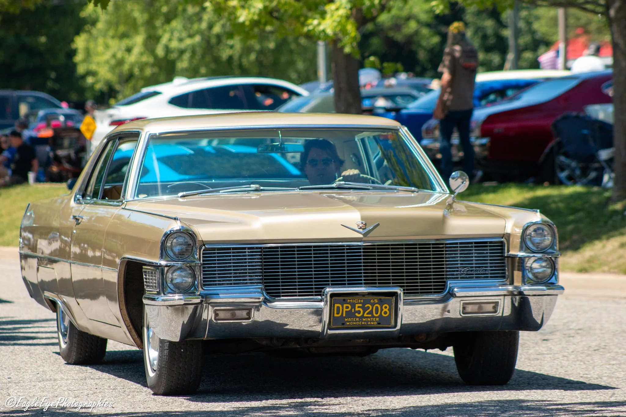 A vintage gold Cadillac parked at an outdoor car show, with a person inside the car and a crowd in the background.