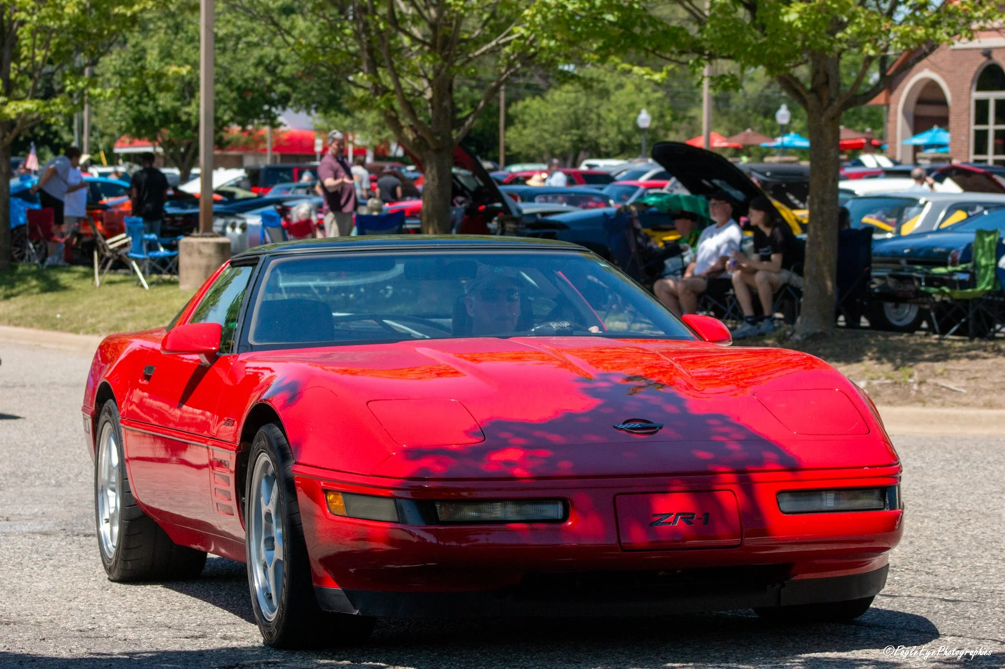 A bright red Chevrolet Corvette ZR-1 sports car with a sleek design is parked on the street at a car show. In the background, there are people sitting and walking among a variety of other colorful cars and trees, indicating an outdoor event on a sunn