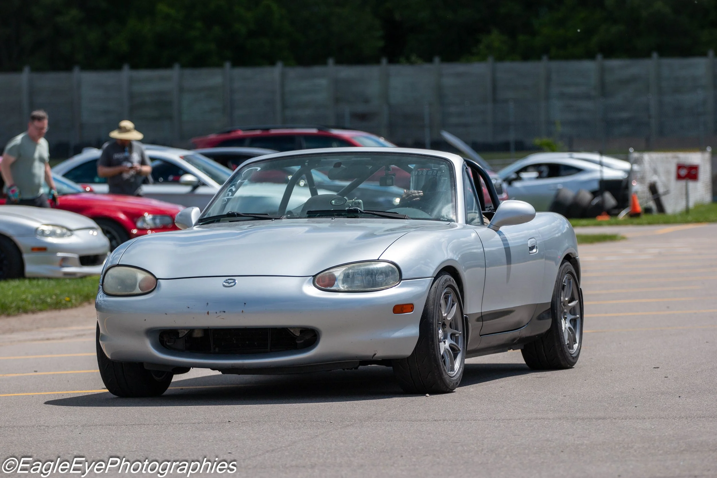 Silver Mazda convertible car parked in an outdoor parking lot during daytime with other cars and people in the background.