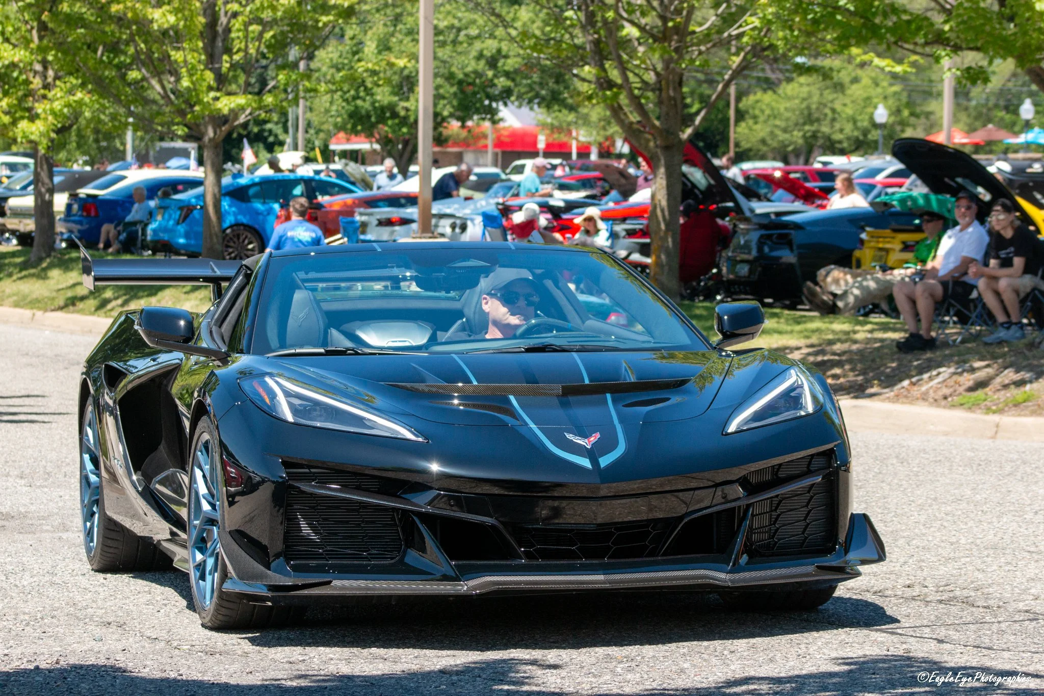 A black Chevrolet Corvette sports car with blue racing stripes in the foreground, at a car show with people and various vehicles under trees in the background.