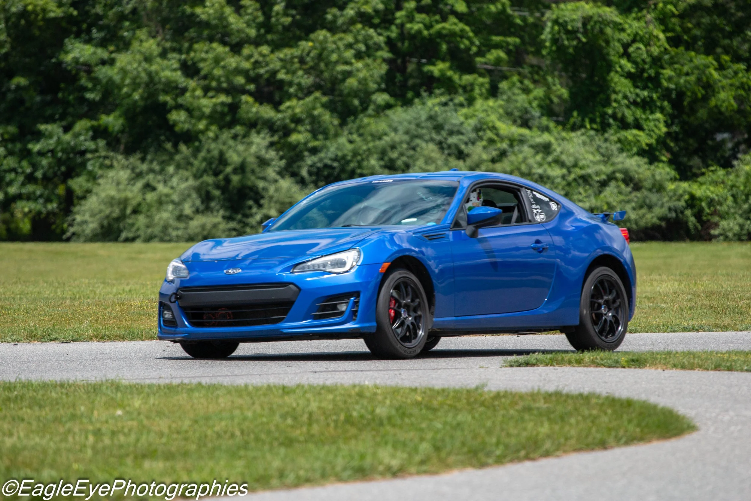 Blue sports car driving on a grassy area with green trees in the background.