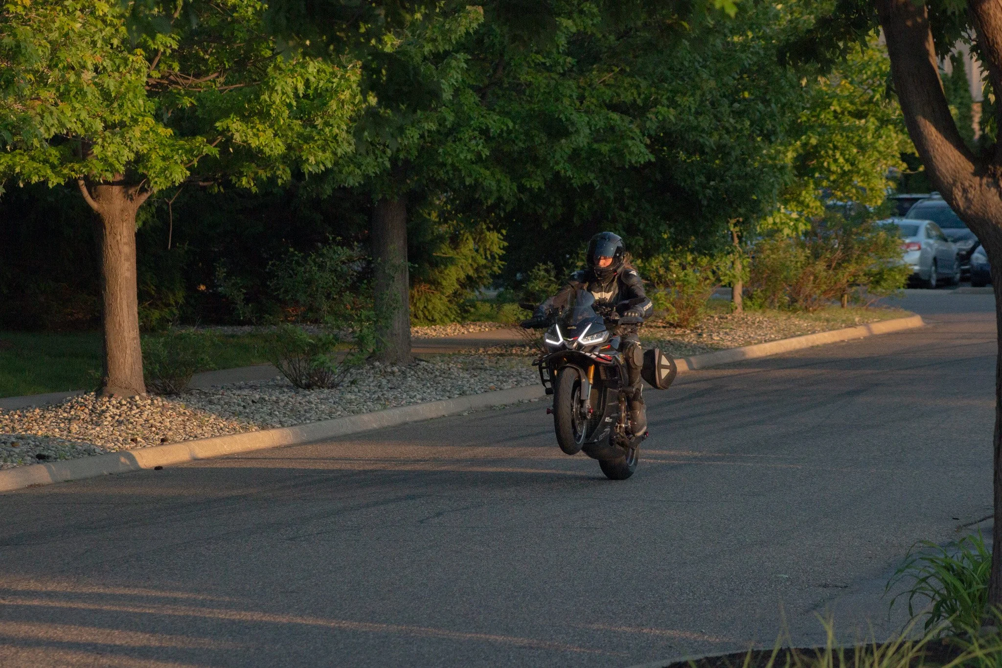 A motorcyclist wearing a black helmet and black riding gear riding a black motorcycle on a paved road near green trees and parked cars.
