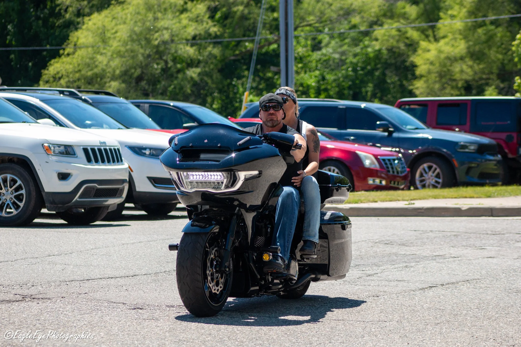 A man riding a black motorcycle in a parking lot with multiple parked cars and green trees in the background.